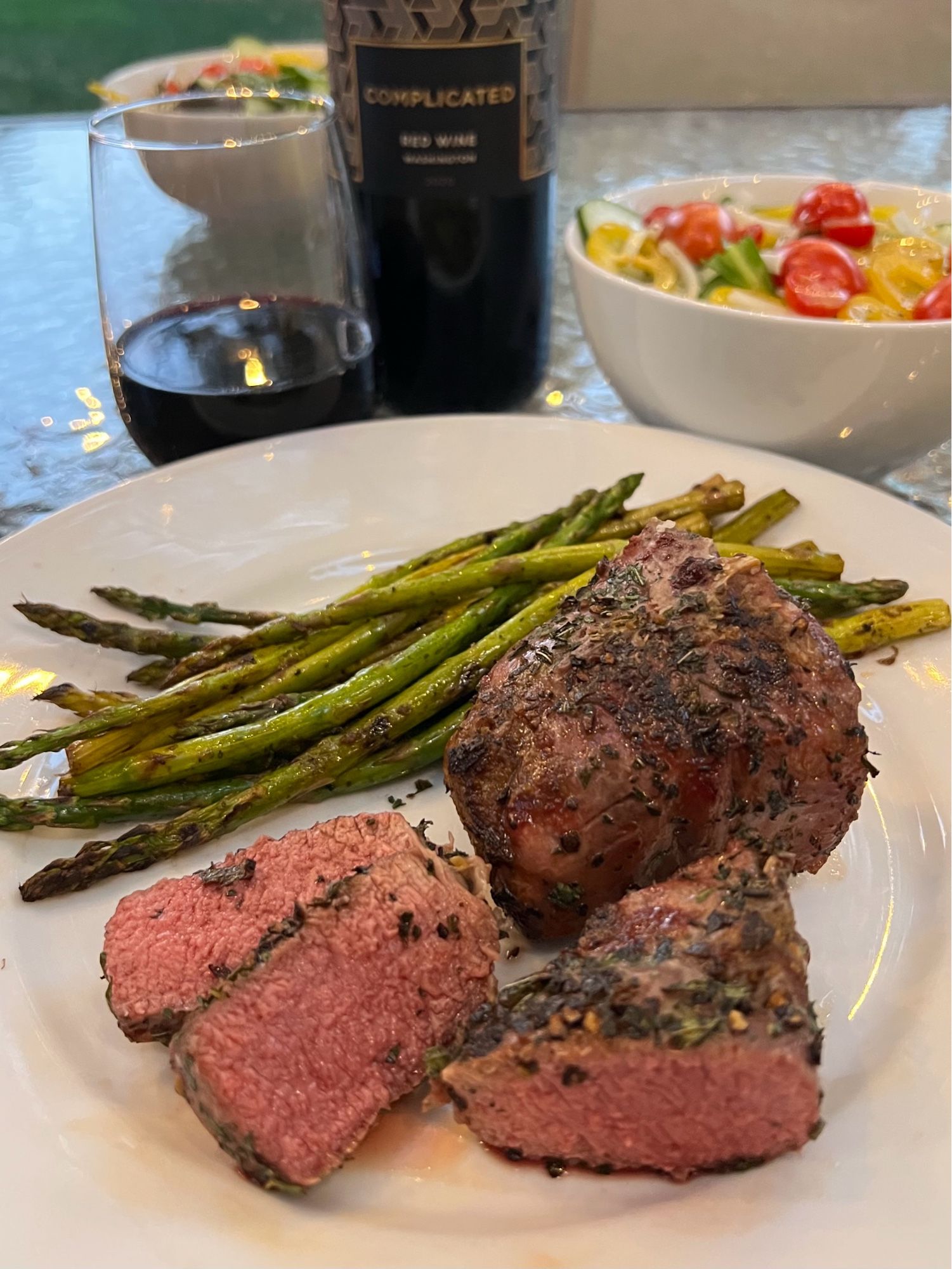 Grilled lamb with rosemary and asparagus with lemon zest, garlic and balsamic vinegar served on a white plate. Small salad with yellow peppers and tomatoes in the background. And a bottle and glass of red wine from Washington.