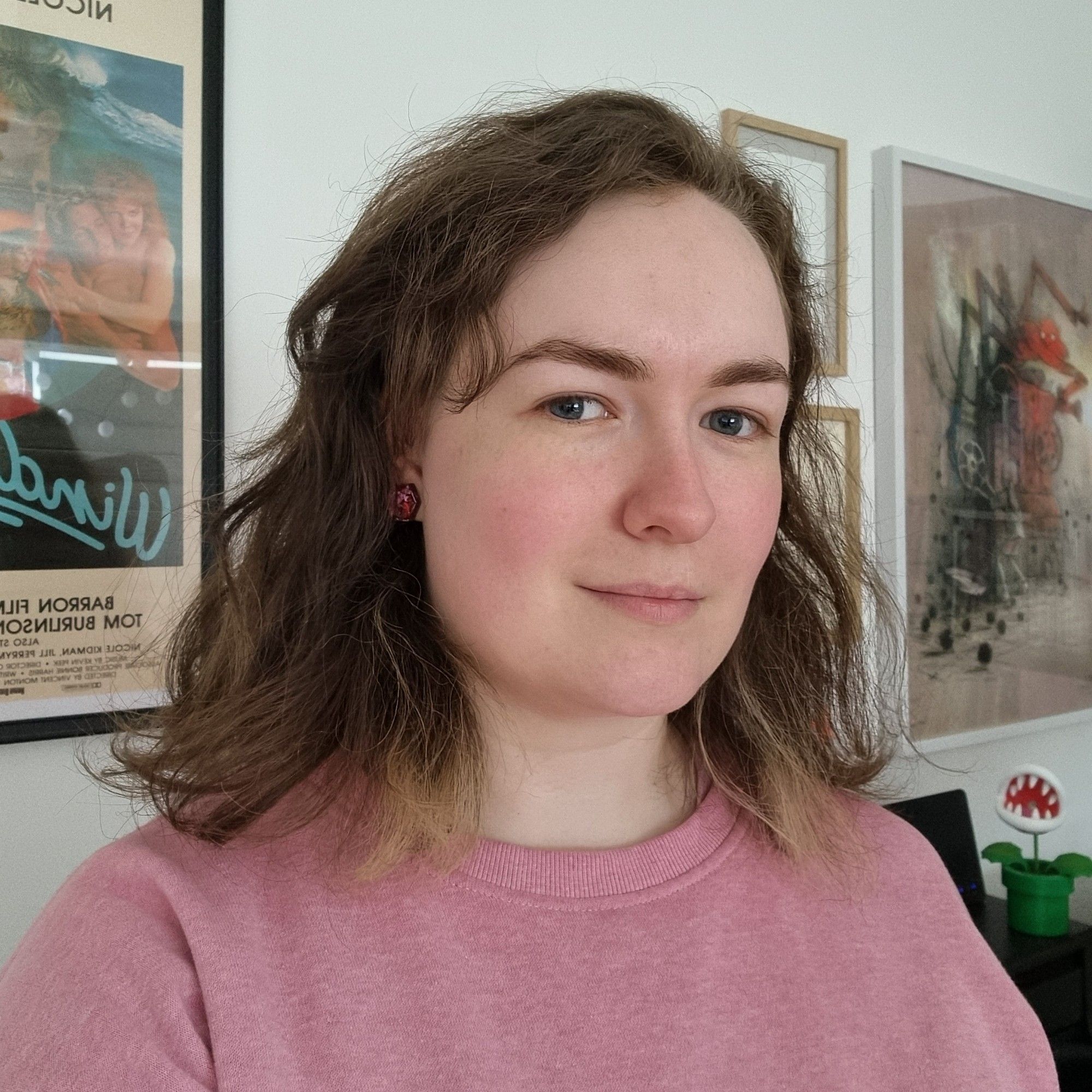 It's me! A brown-haired white woman wearing a pink jumper smiles at the camera. Behind me there's posters for Windrider and Shaun Tan's commemorative release for a Madman Studio Ghibli event in 2017.