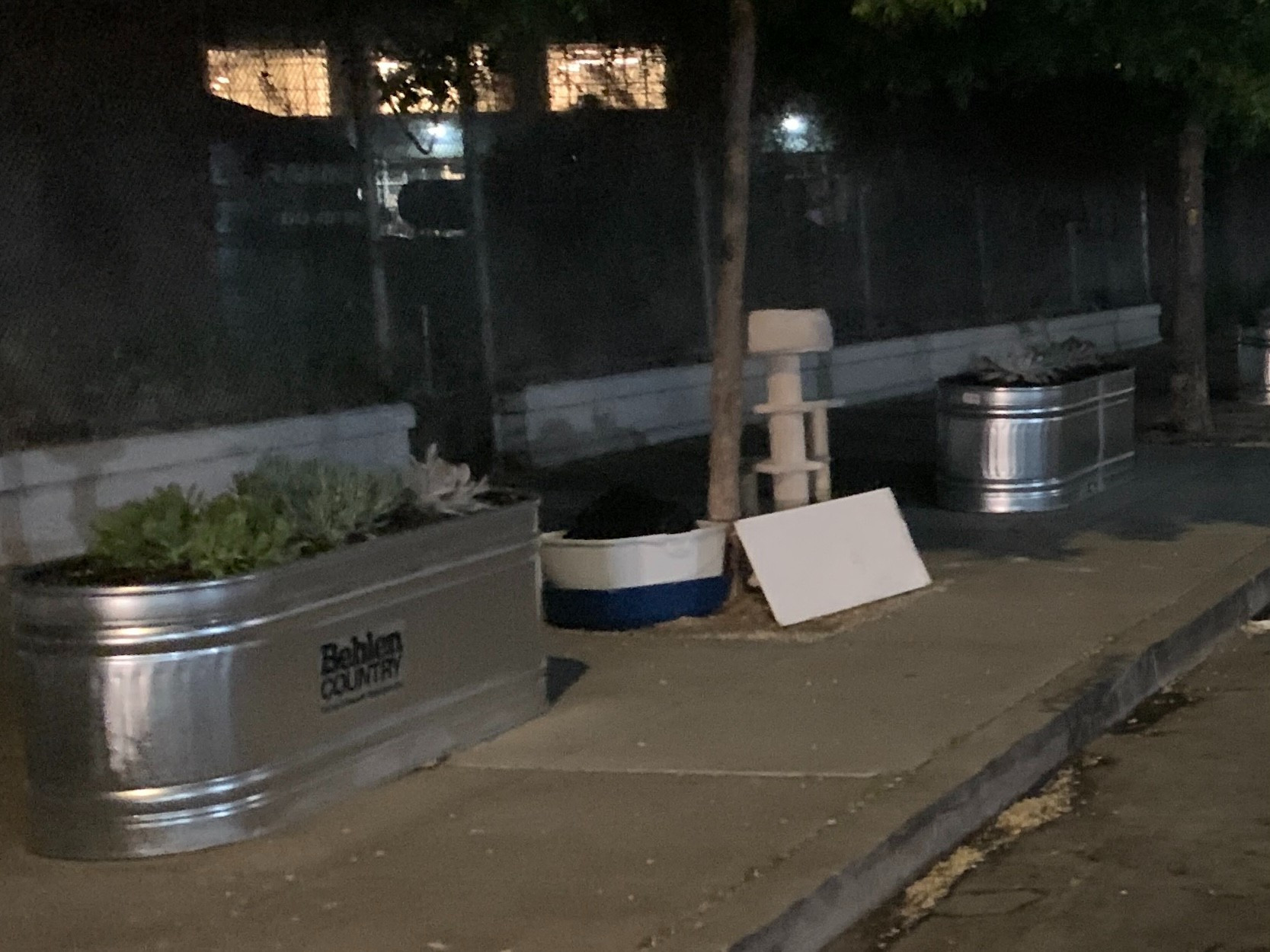 A 6 ft. metal planter full of dirt and plants on a public sidewalk. It's near a parking lot, but on the regular sidewalk. Next to the planter is some trash like a cat tree and empty buckets.