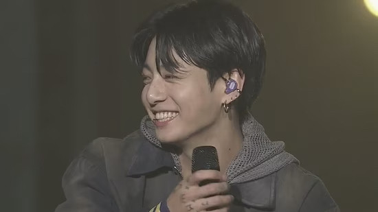 Close up of Jungkook on stage. He’s holding a microphone, wearing a grey hoodie and jacket. We can see his purple in-ear. He’s looking to the side with a huge smile on his face.