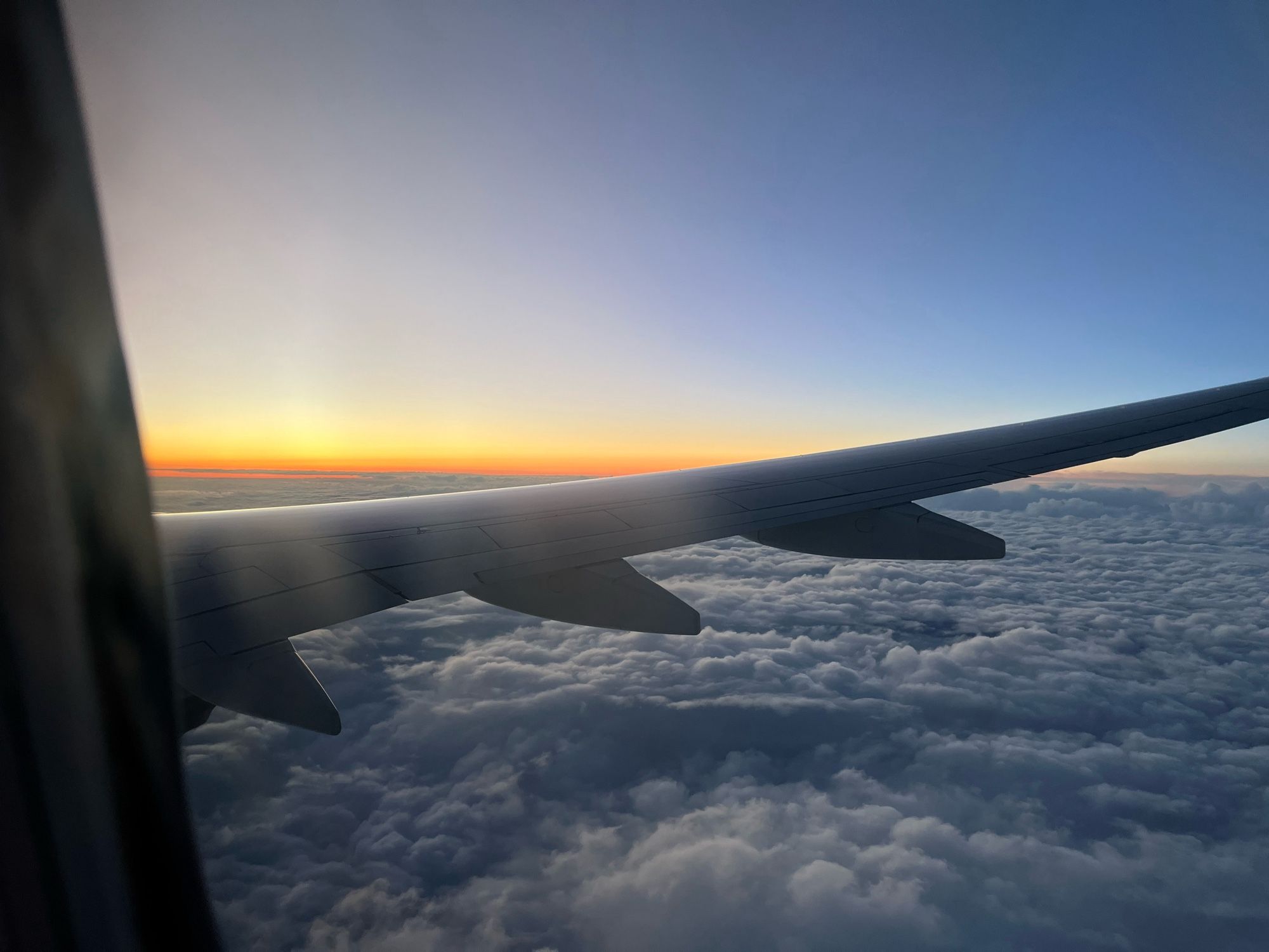 Sunset from the window of an airplane. Yellow and orange above a layer of fluffy clouds. The plane’s wing bisects the photo.