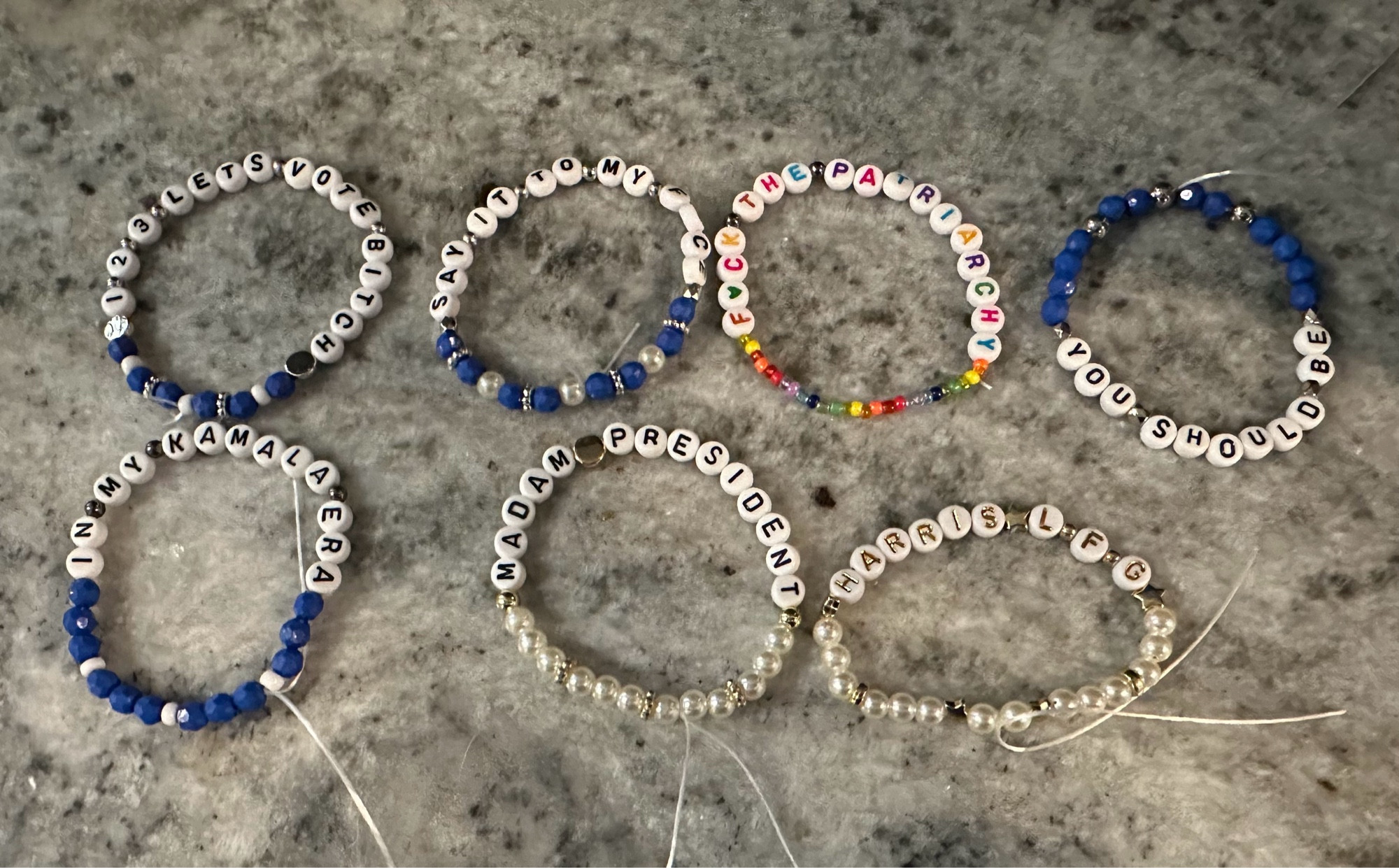 Seven beaded bracelets with various colors and phrases on a gray surface.

Text Transcription:

1 2 3 LETS VOTE BITCH
SAY IT TO MY FACE
F*CK THE PATRIARCHY
IN MY KAMALA ERA
MADAM PRESIDENT
YOU SHOULD BE
HARRIS LFG