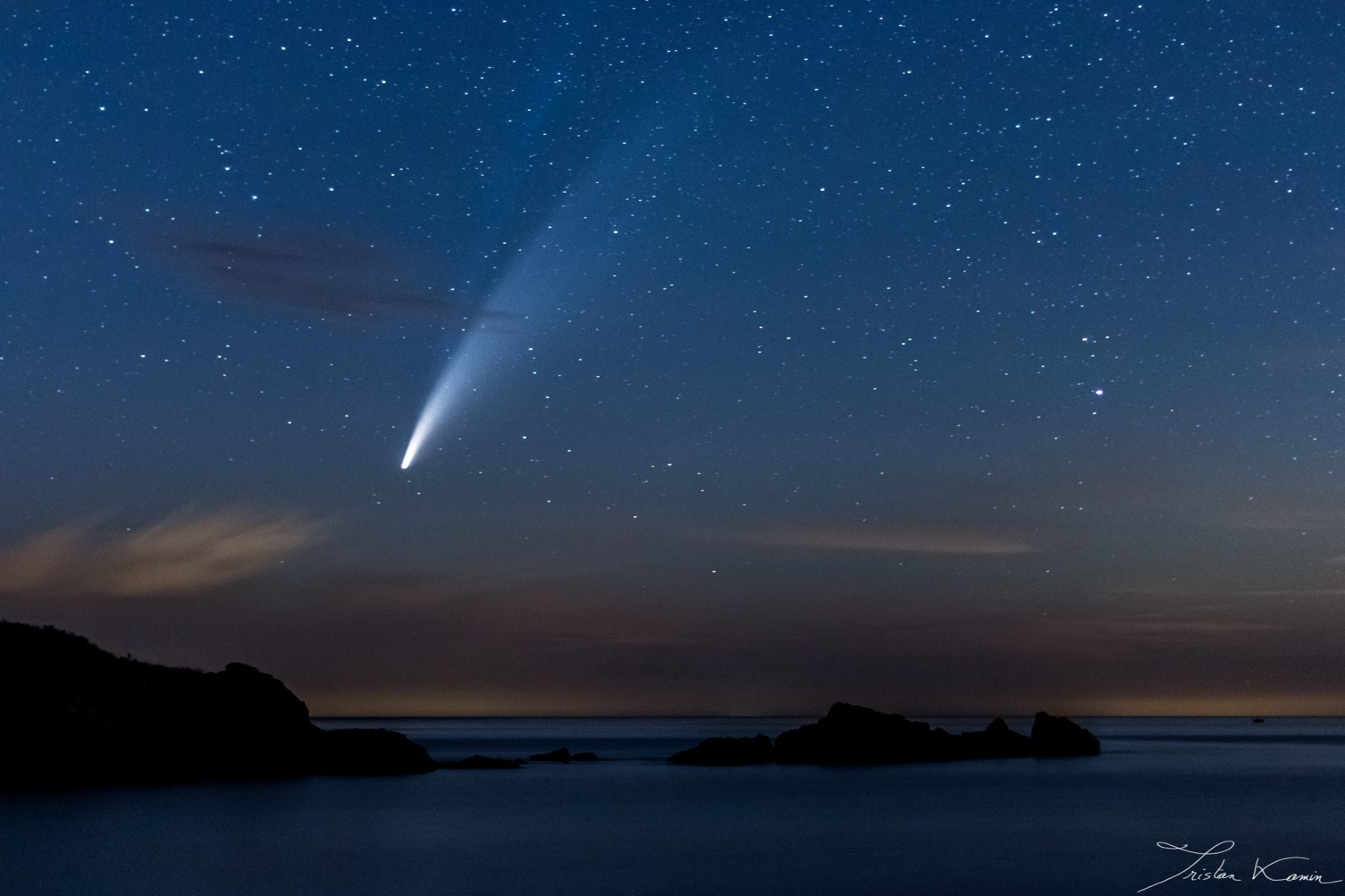 Photographie du bord et mer, l'eau lissée par le temps de pose est découpée par la silhouette de rochers émergents. Le ciel est chargé de nuages orangés proches de l'horizon mais, au-dessus des nuages, au milieu d'un ciel riche en étoiles, une imposante comète déchire le ciel. La deuxième queue de la comète est légèrement distinguable.