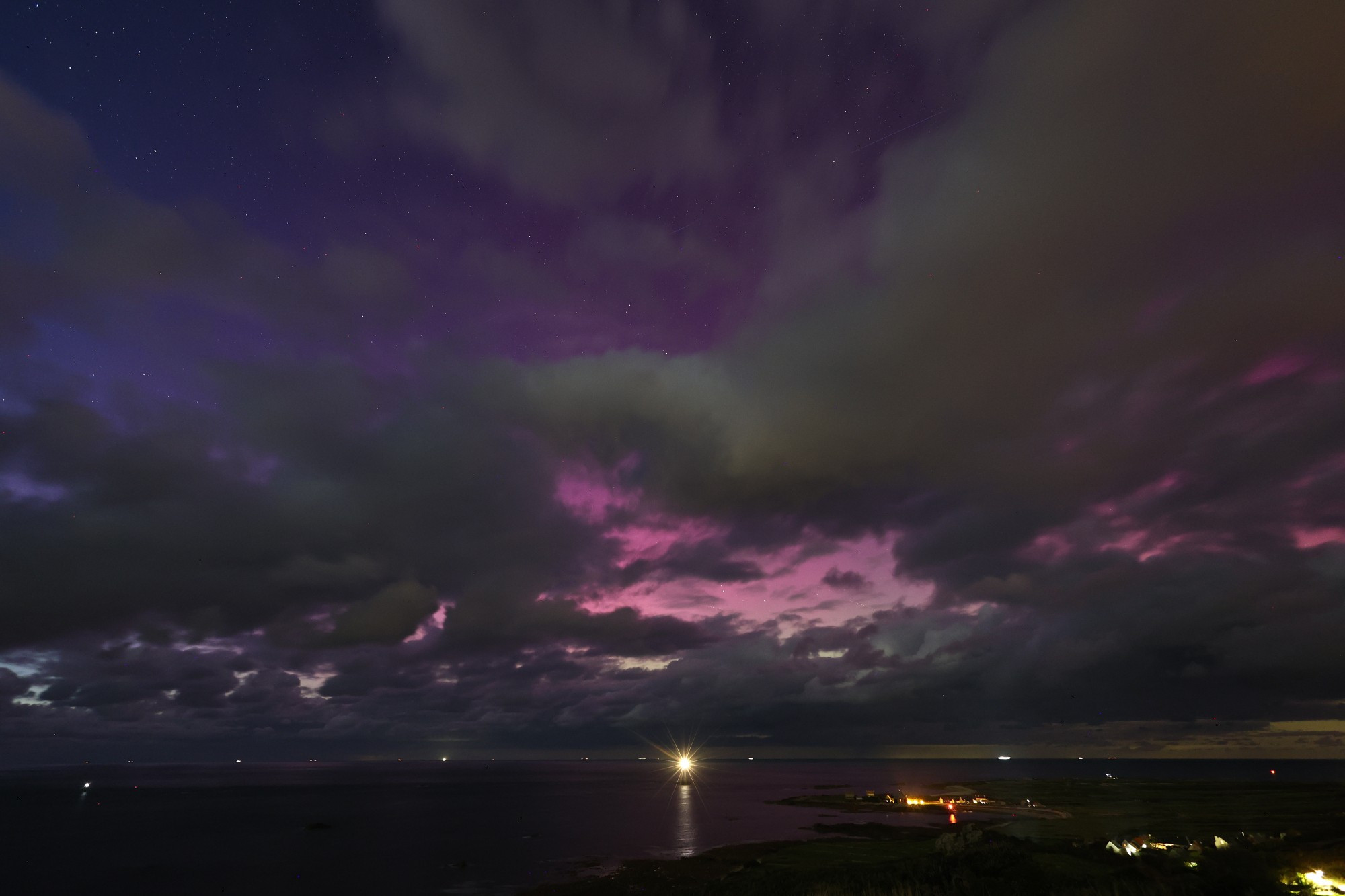 Photographie d'une aurore boréale. le ciel est parsemé de nuages fragmentés et élongés mais le gradient de bleu du ciel nocturne au rose pale de l'aurore boréale, avec tout le nuancier de rose et de violet est bien présent. À l'avant plan, une étendue d'eau, un peu de terre qui s'avance en presqu'île dans l'eau et un phare.