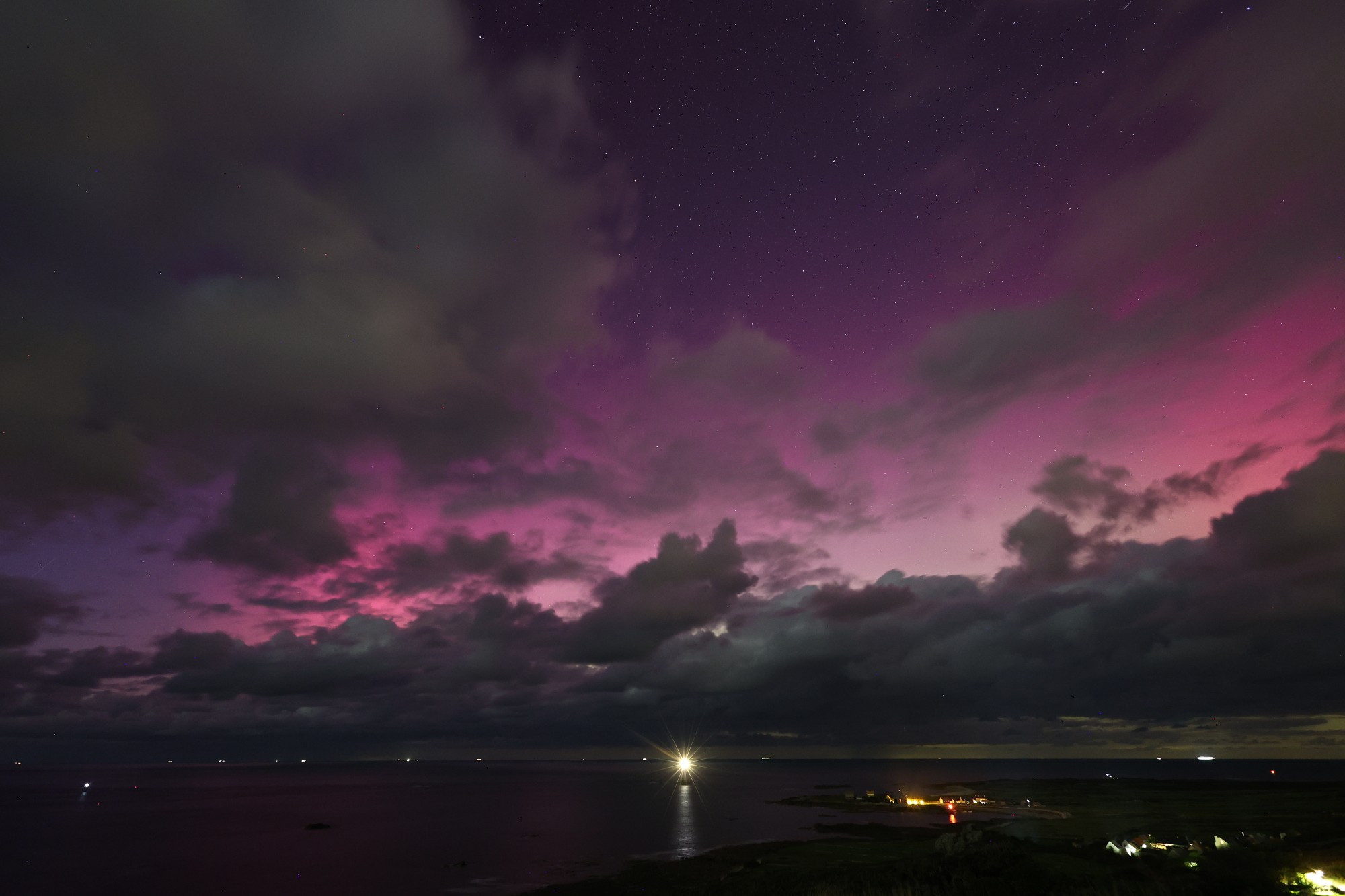 Photographie d'une aurore boréale. le ciel est parsemé de nuages fragmentés mais le gradient de violet au rose pale de l'aurore boréale est bien présent. À l'avant plan, une étendue d'eau, un peu de terre qui s'avance en presqu'île dans l'eau et un phare.