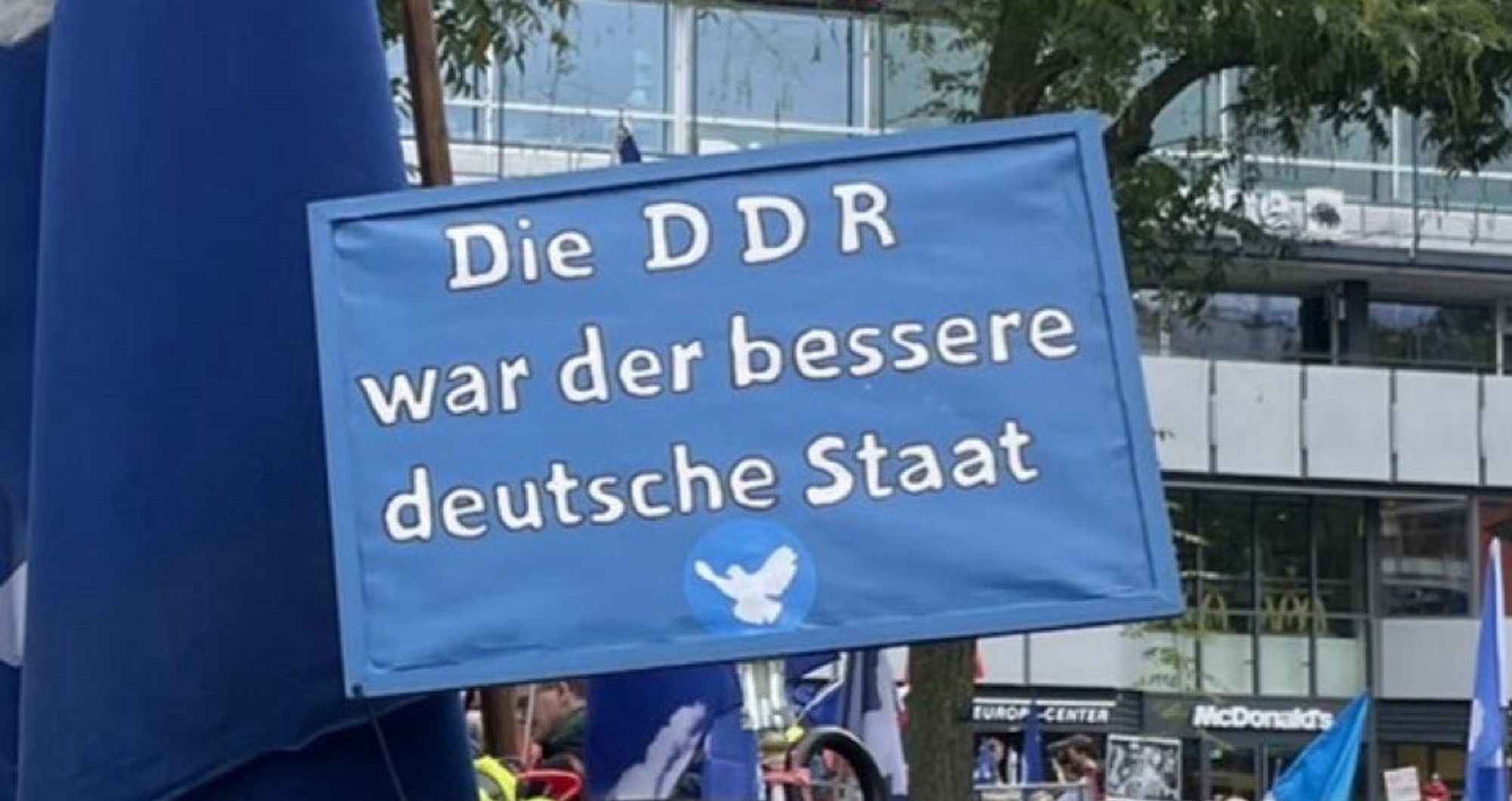 Zu sehen ist ein blaues Transparent mit einer „Friedenstaube“ auf der heutigen Demo in Berlin. Auf dem steht: „Die DDR war der bessere deutsche Staat“