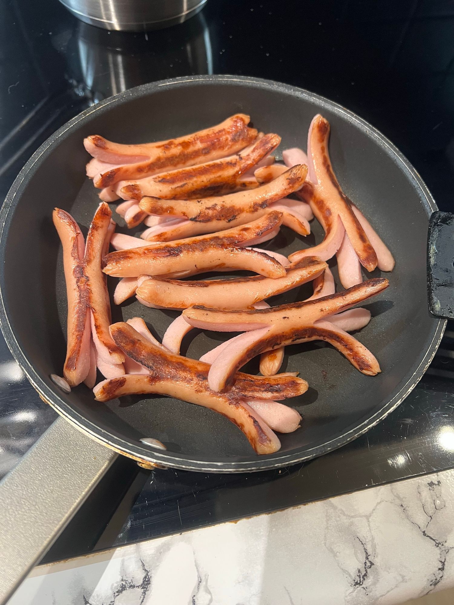 A frying pan containing several browned hot dogs, arranged in a casual layout, on a dark countertop.