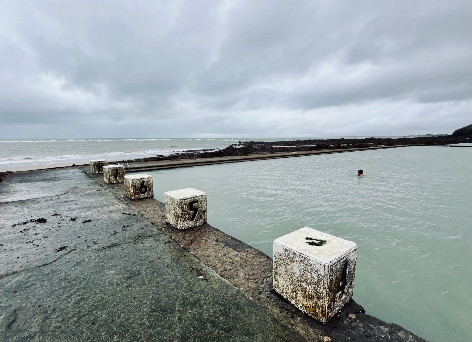 atlantik bei ebbe im hintergrund, im vordergrund ein beton-schwimmbecken mit meerwasser, alte verwitterte sprungblöcke und eine einzelne person mit badekappe im wasser. dramatischer wolkenhimmel.