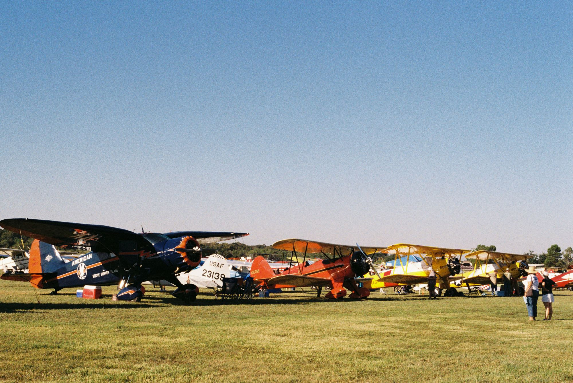 Three vintage planes on a grass field with two people looking at them.