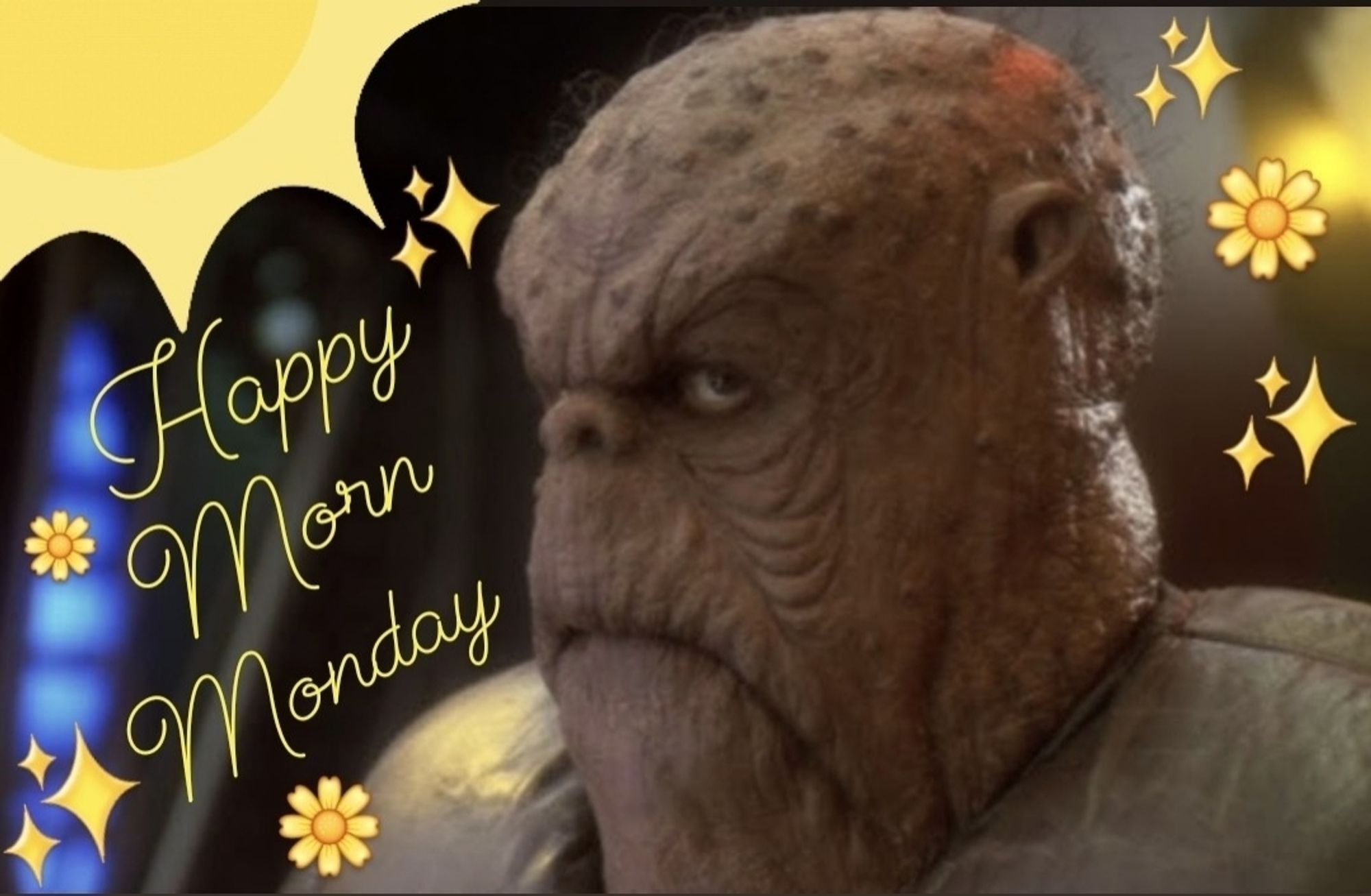 Close up of Morn from Star Trek DS9 Deep Space 9, with his usual, impassive expression. A cartoony yellow sun has been edited into the top left corner of the image, and underneath in a yellow, script font it says: "Happy Morn Monday." Yellow sparkle and flower emojis are scattered about, decorating the rest of the image, giving it a cheery, summery, morning vibe.