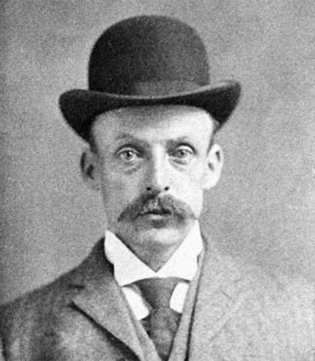 A blavk and white photo of middle-aged white man with a large mustache. He is wearing a suit and bowler hat, and stares blankly at the camera.