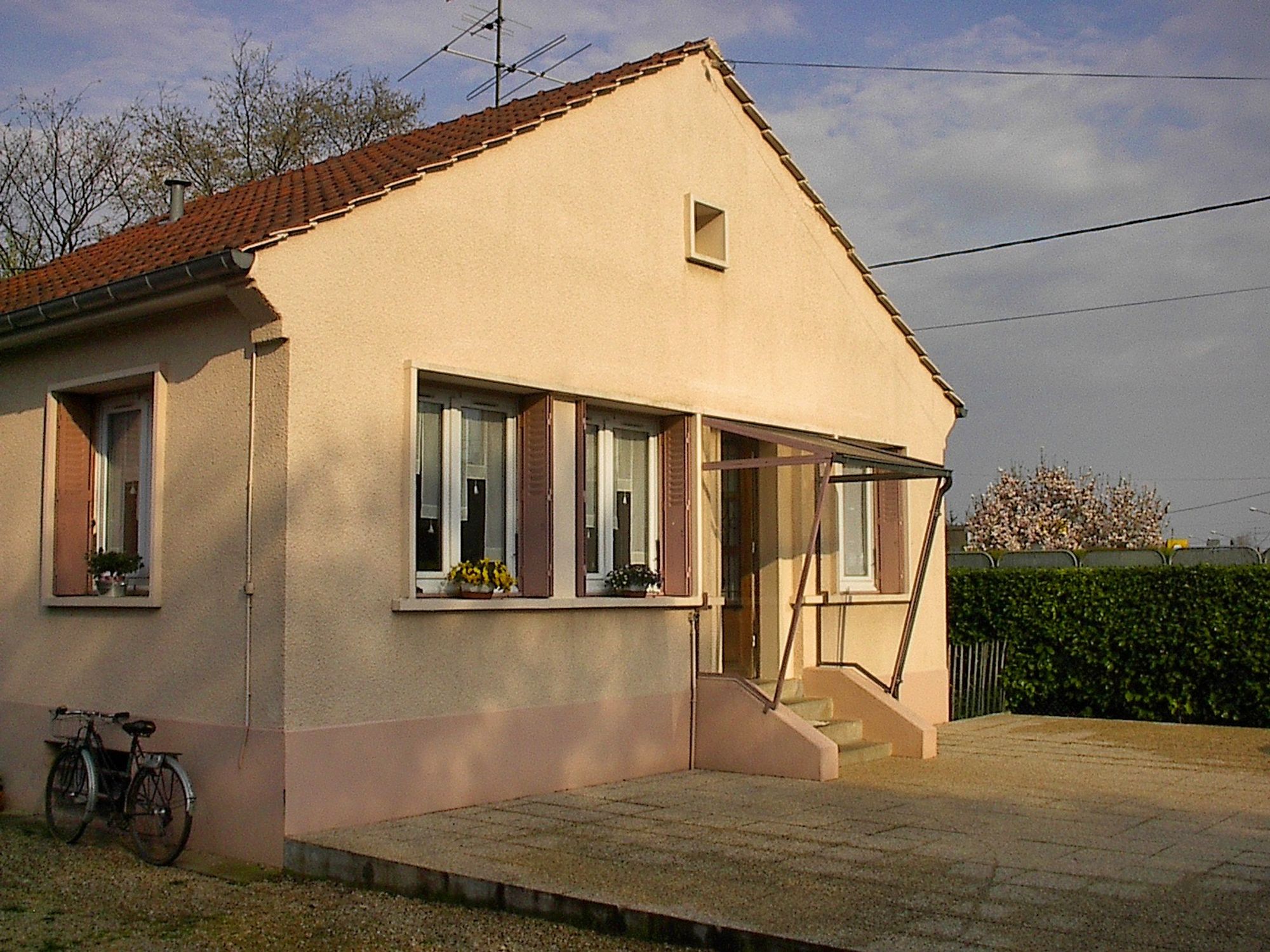 Photo d'une maison prise sans être en face. Des magnolias en fleur au loin et les couleurs laissent penser qu'elle a été prise à l'heure dorée un matin ou un soir de printemps. En façade, 3 fenêtres (dont deux fleuries) et un petit perron avec 4 marches d'escalier. Un vélo est posé contre le mur sur le coté. Devant, une terrasse, et du coté où se trouve le vélo, une allée en gravier.