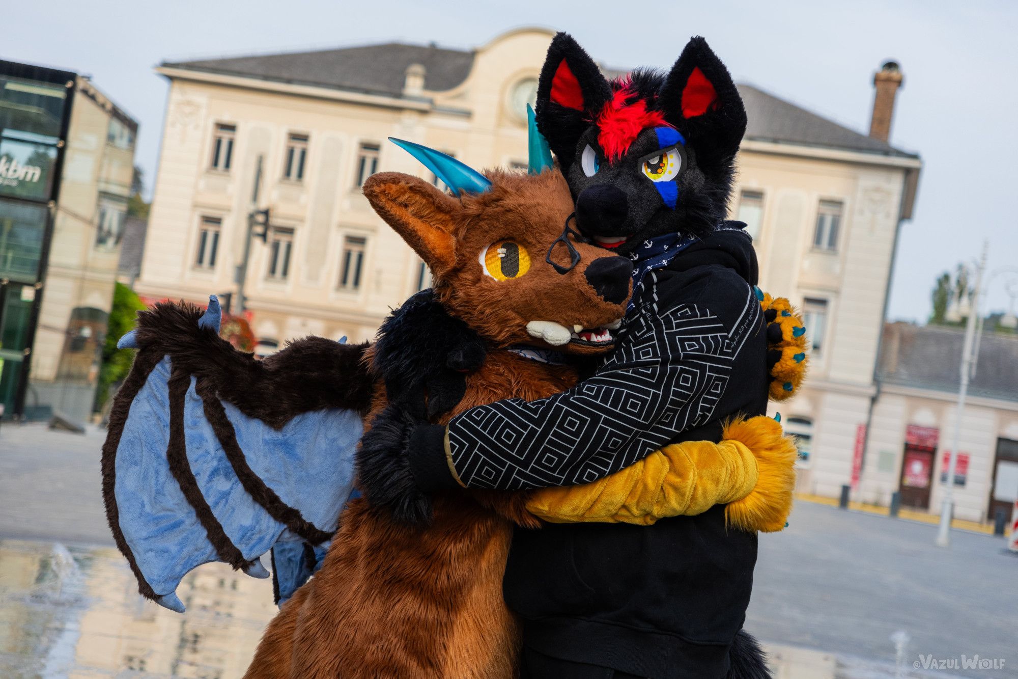 A picture showing two fursuiters hugging each other. Rayu, a brown furred, winged fursuiter is on the left and hugging a black fox fursuiter. Both suiters are standing sideways, which makes Rayu's wings visible