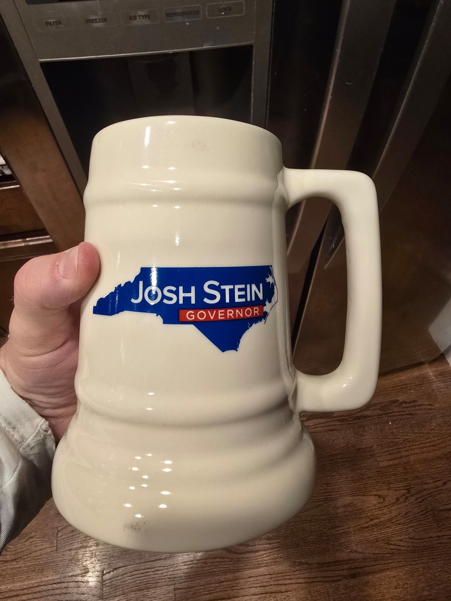 This is a beer stein. It overflows with the promise of beer-related campaign merchandise. The stein advertises Josh Stein, attorney general of North Carolina, for governor.  The name is in a blue outline of North Carolina. The stein is white polished ceramic