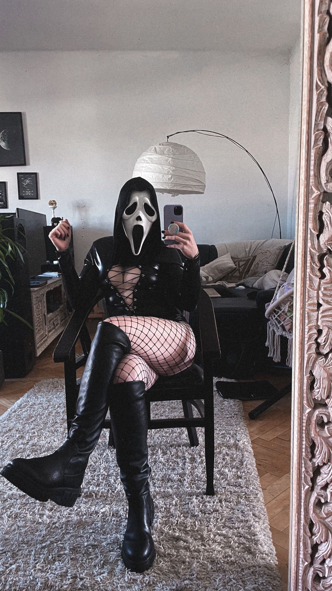 girl sitting on a dark brown chair in front of her mirror, wearing a leathersuit, fishnets and black boots combined with a ghostface mask