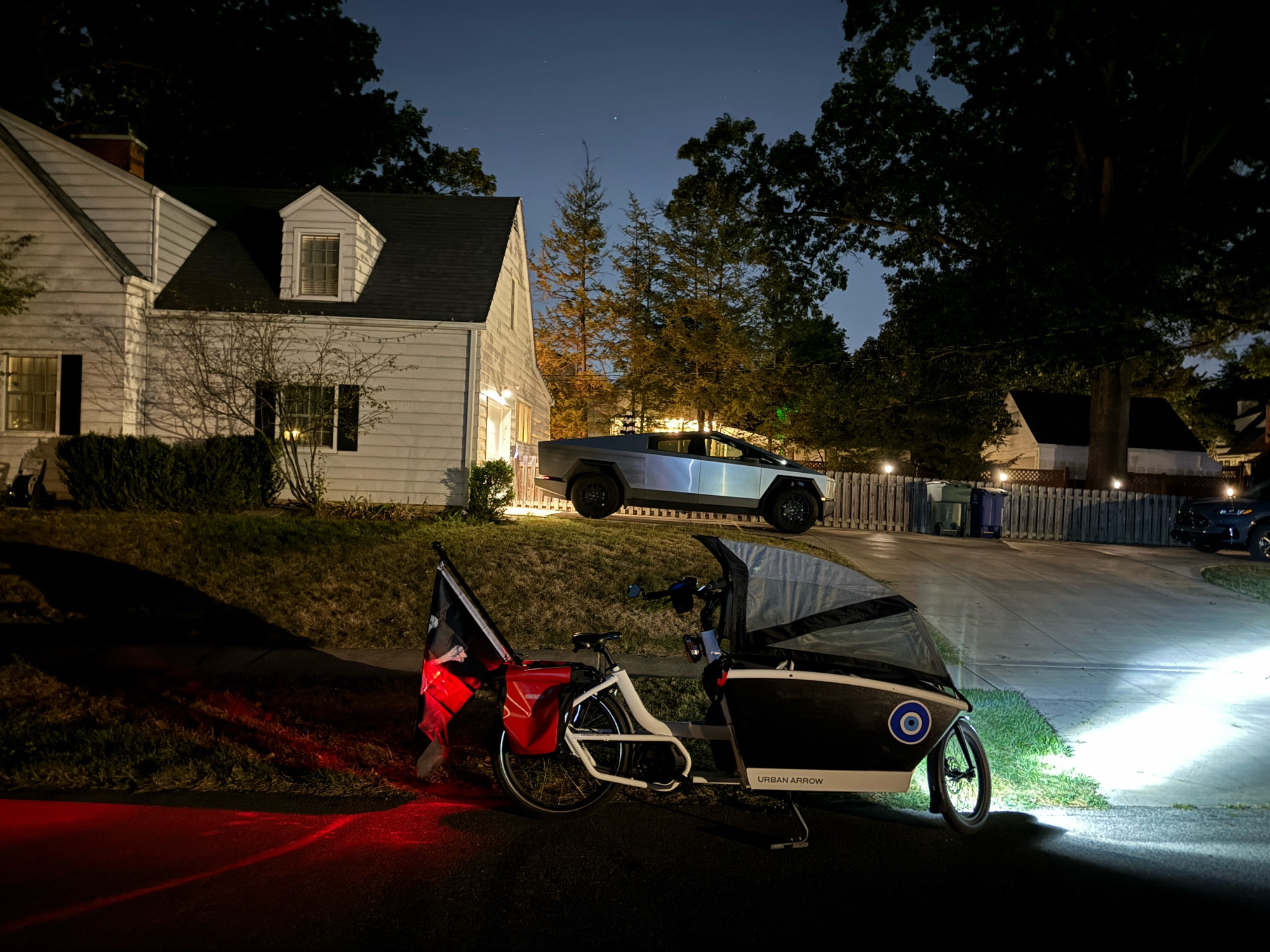 A Tesla Cybertruck in the background in a nighttime photograph. An Urban Arrow with lights lit, flying a pirate flag from the rear tail, in the foreground.