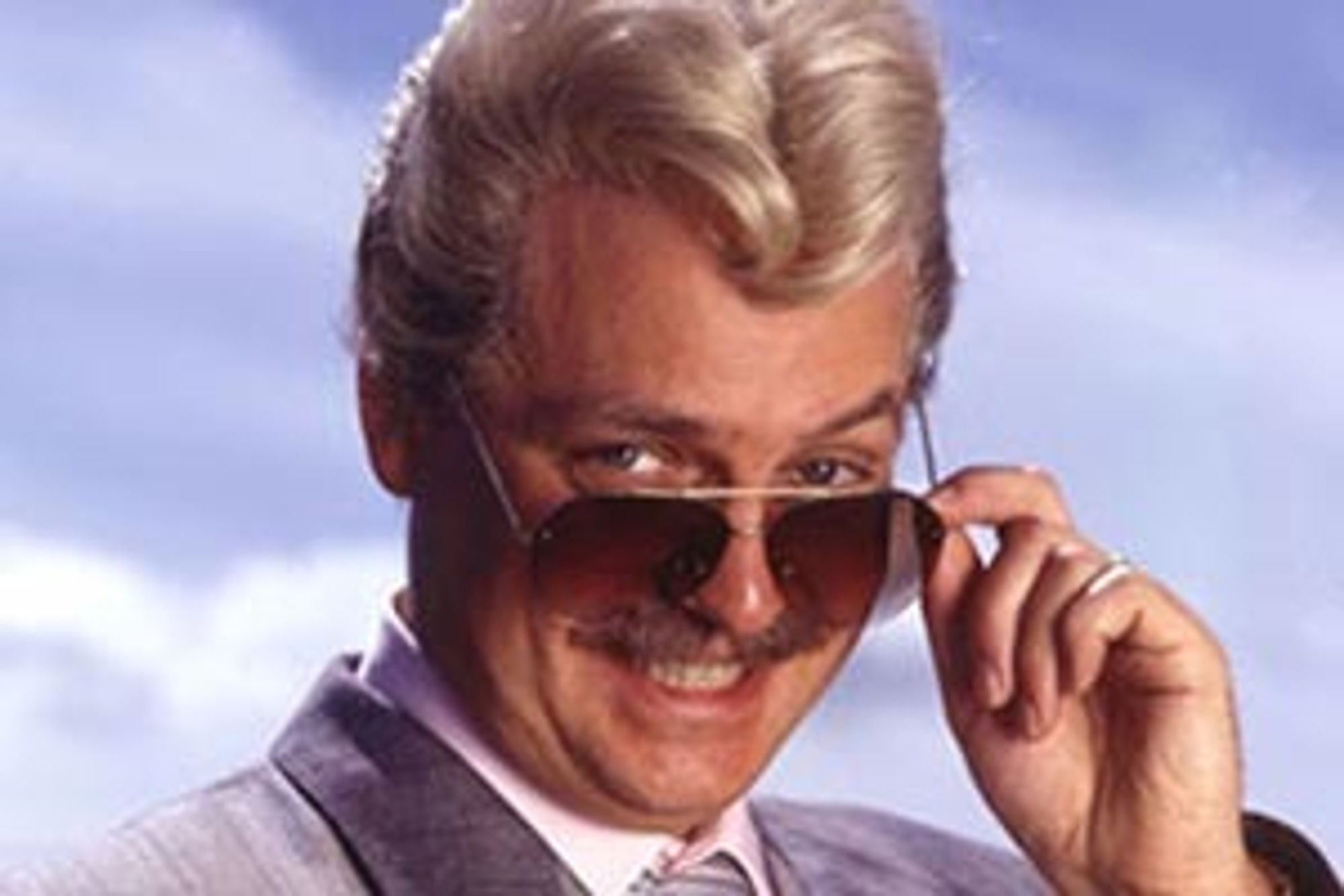 A photograph of the character Swiss Tony from "The Fast Show" - a sleazy man with a silver quiff hairdo grinning lecherously over the top of his Ray-Ban sunglasses.