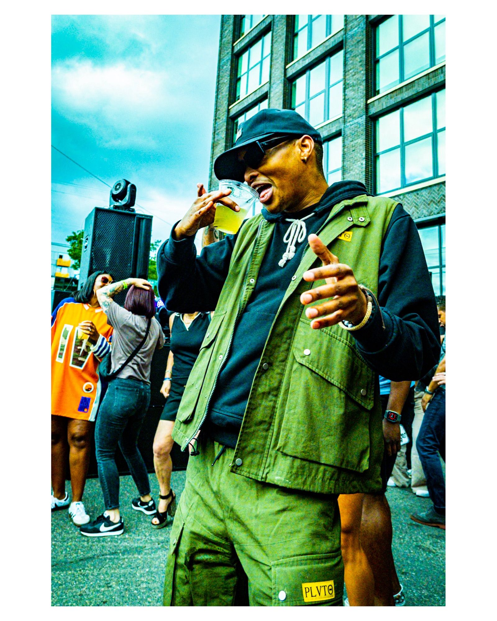 Young Black man in matching green shorts and vest (worn over a hoodie), black glasses and hat, sipping on a cocktail while dancing to house music at a block party. Background is other dancers, the partly cloudy sky and the brick and glass facade of the adjacent building.