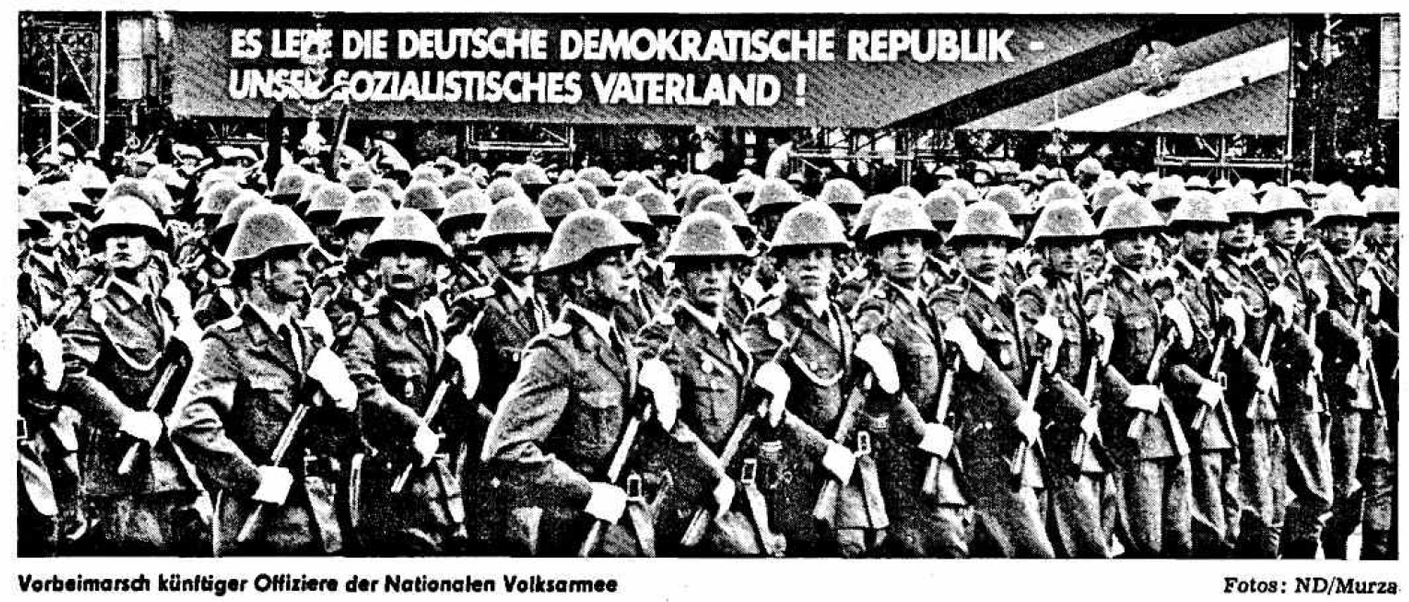 Soldaten mit Stahlhelm und Gewehr marschieren in Formation vor einem Plakat:
Es lebe die deutsche demokratische Republik - unser sozialistisches Vaterland!
Unterschrift: Vorbeimarsch künftiger Offiziere der Nationalen Volksarmee