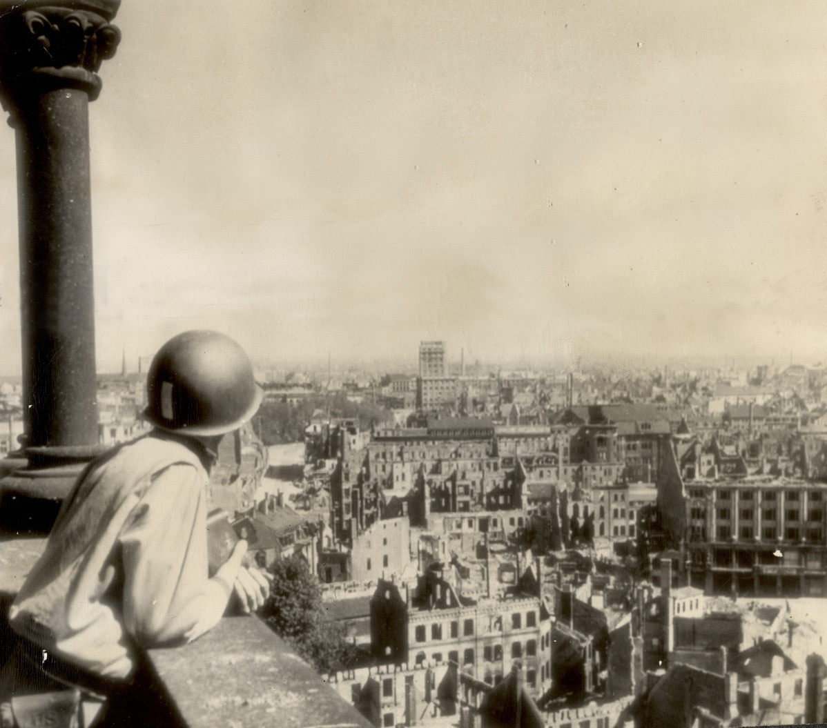 Ein Soldat mit Stahlhelm lehnt sich über eine steinerne Brüstung vor ihm liegen zerstörte Häuser bis zum Horizont neben ihm eine Säule