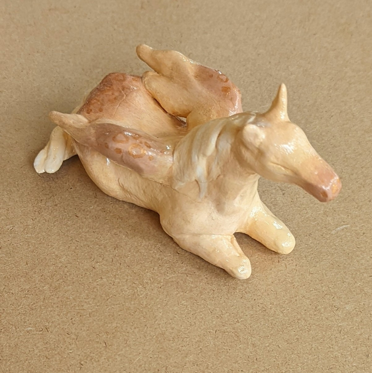 "Pegasus Foal", 6" by 2.5" by 3" natural clay sculpture depicting a cream- and gold- colored pegasus resting