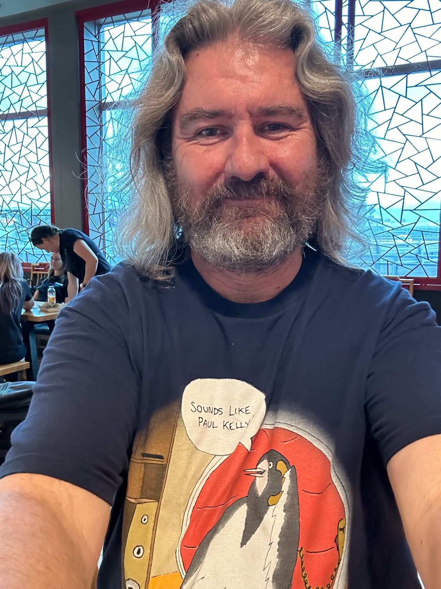 Me, wearing a navy T-shirt with a picture of a magpie-lark in a chair sitting near a speaker, holding a phone and sayi g “Sounds like Paul Kelly!”