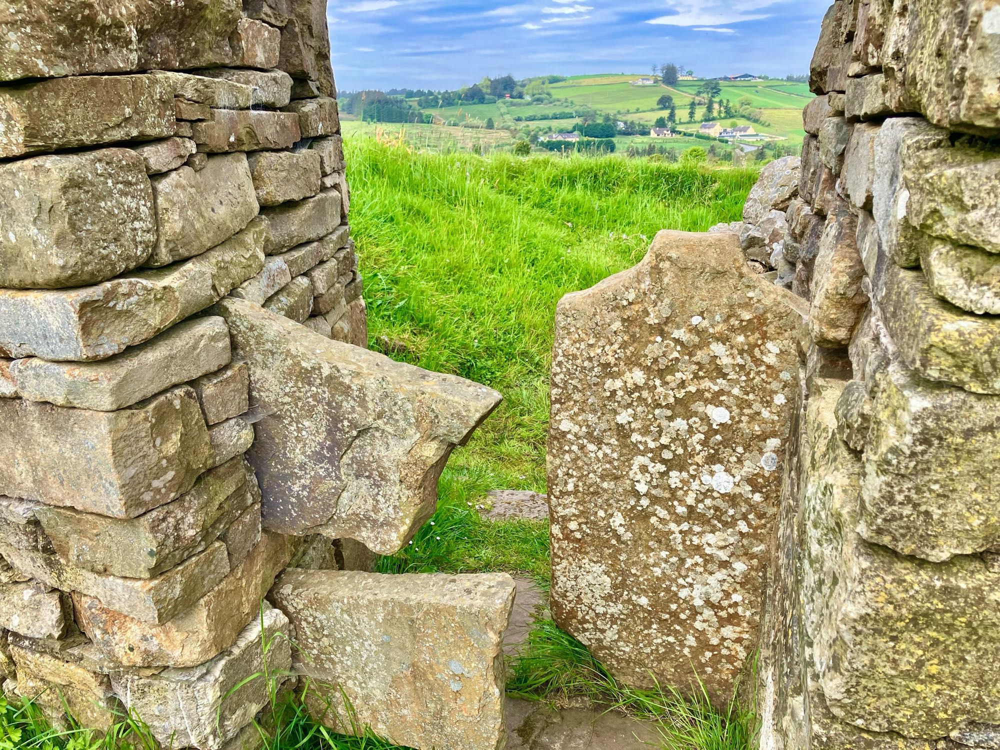 A stone entrance to an ancient Irish holy well with blue skies and sunshine on distant fields at Brosna in county Kerry