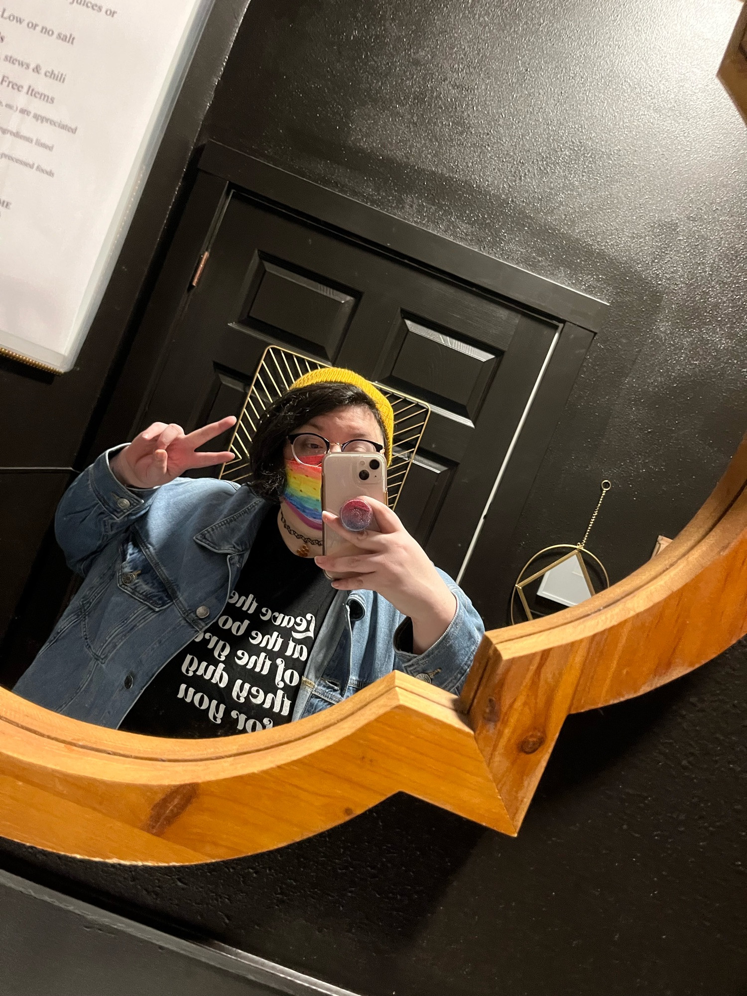 a mirror selfie of quinn. they wear a yellow beanie, a rainbow mask, a denim jacket, and a black t-shirt with white text on it. they hold their phone in one hand while making a peace sign with the other. 