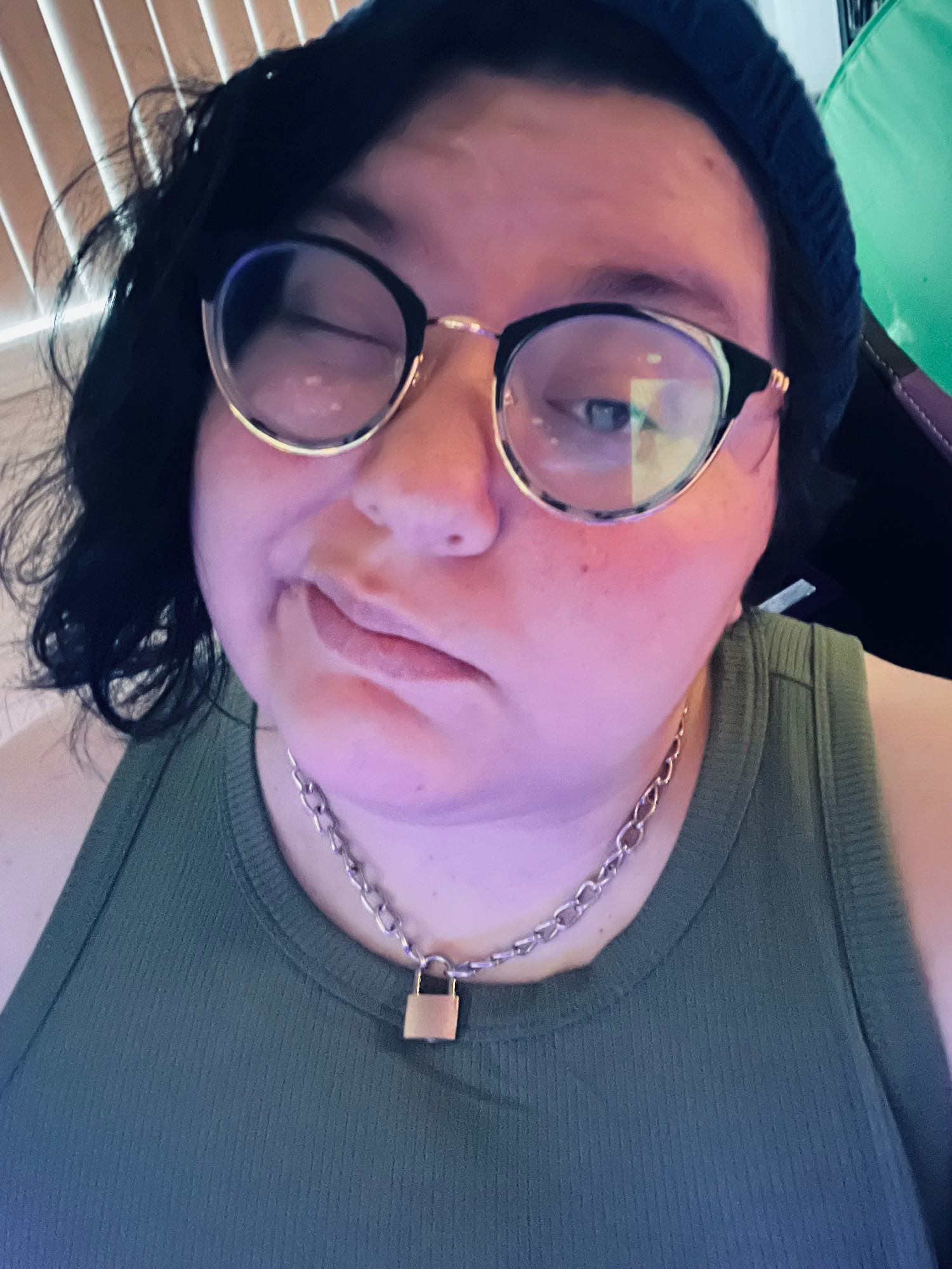 a selfie of quinn. they wear a navy beanie, an olive green tank top, and a silver chain necklace with a bronze padlock on it. 