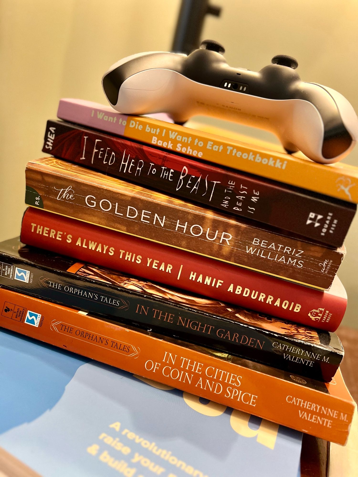 stack of books and a PS5 controller on an end table. mostly fiction, two nonfiction.