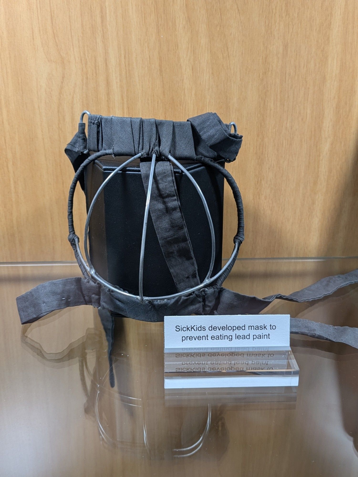 A wire muzzle in a display case. The label says it is a mask developed at SickKids hospital to prevent eating lead paint.