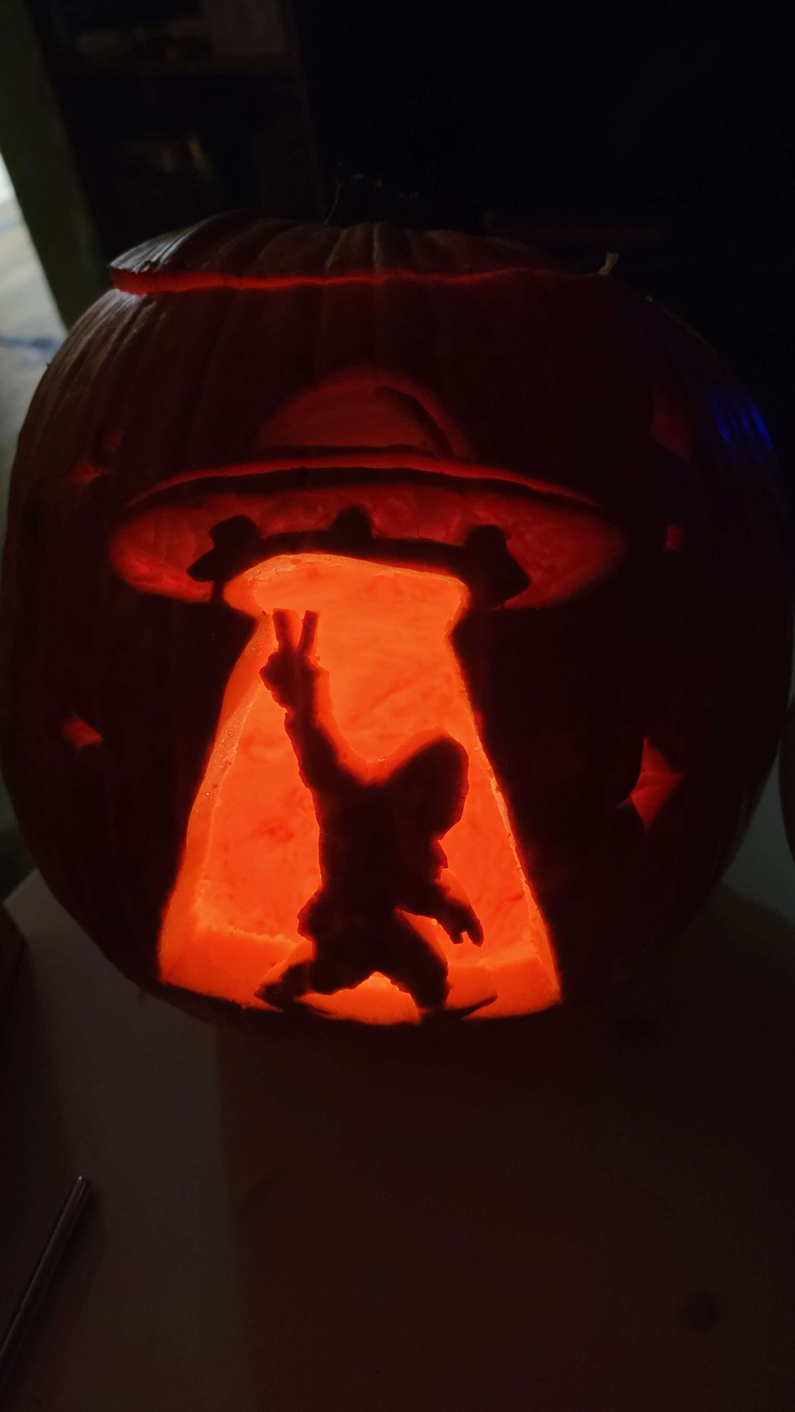 A pumpkin with a UFO carved out and Sasquatch in the beam throwing a peace sign but in the dark to show the candlelight showing through.