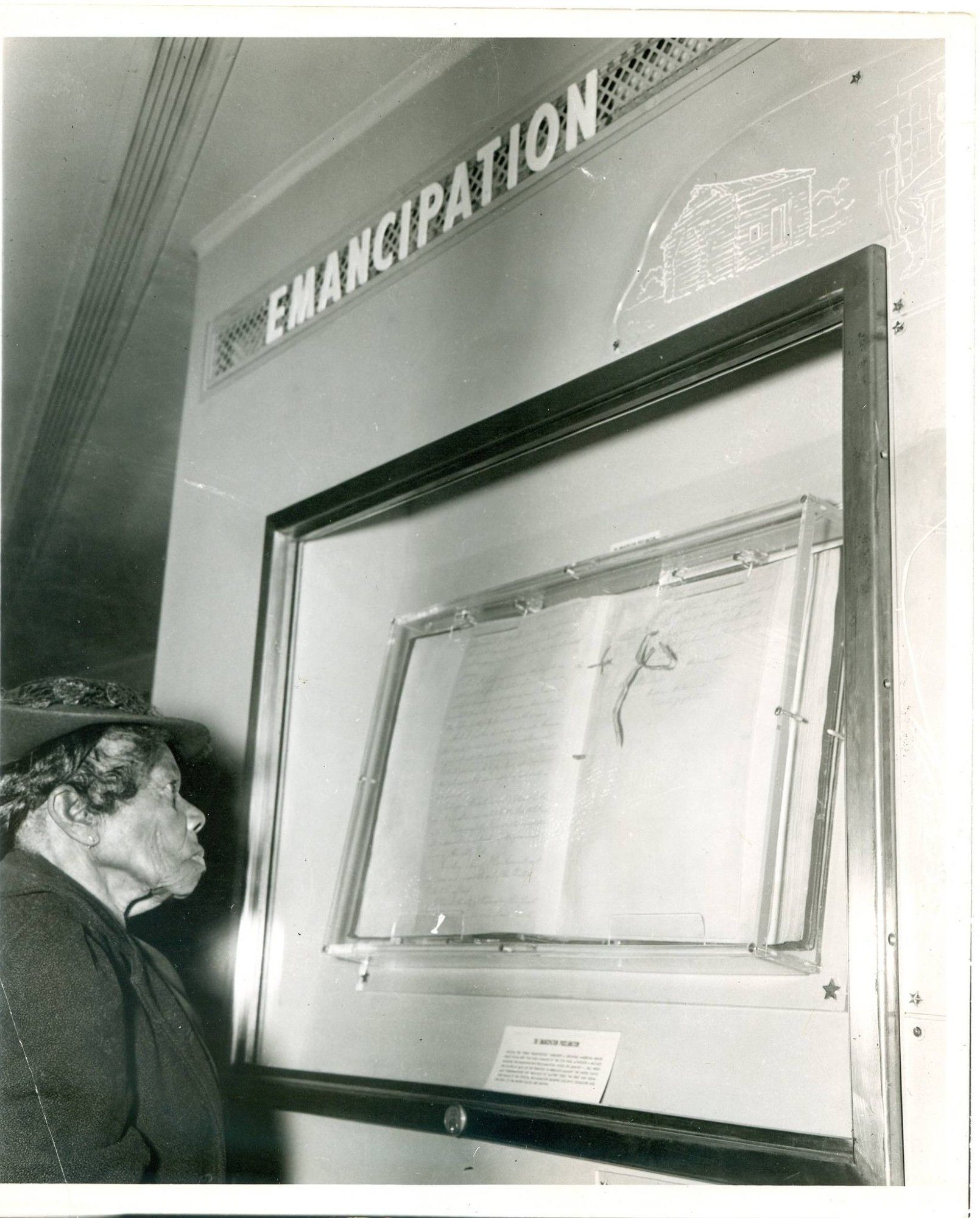 An elderly African American woman wearing a dark coat and hat is pictured on the left. She is reading the Emancipation Proclamation which is displayed in front of her behind glass. Above it in bold white letters is a display that reads EMANCIPATION.