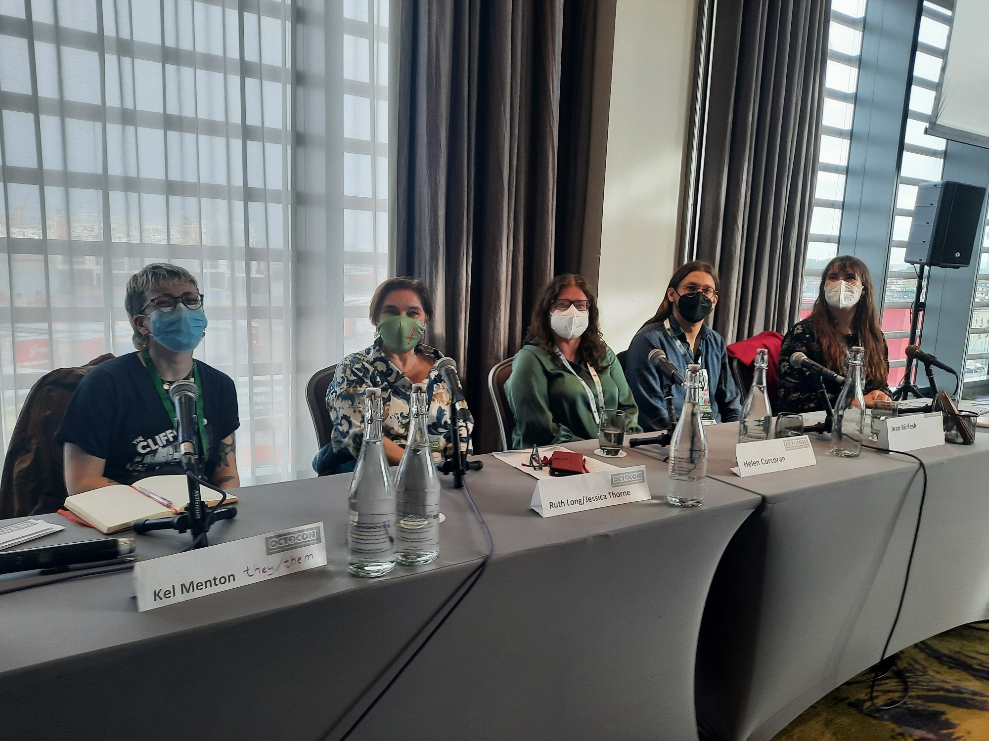 The photo includes Kel Menton, Ruth Frances Long, Helen Corcoran, Jean Bürlesk, and Sarah Rees Brennan sitting on a long table with grey tablecloth wearing face masks