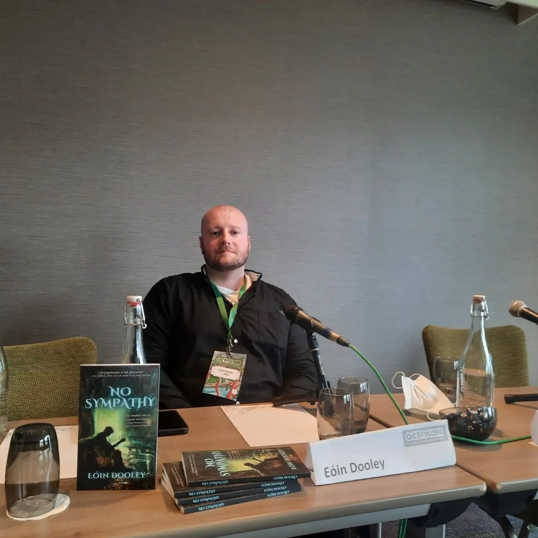 Eoin Dooley sits at a table with a microphone, copies of his book No Sympathy and a glass water bottle.