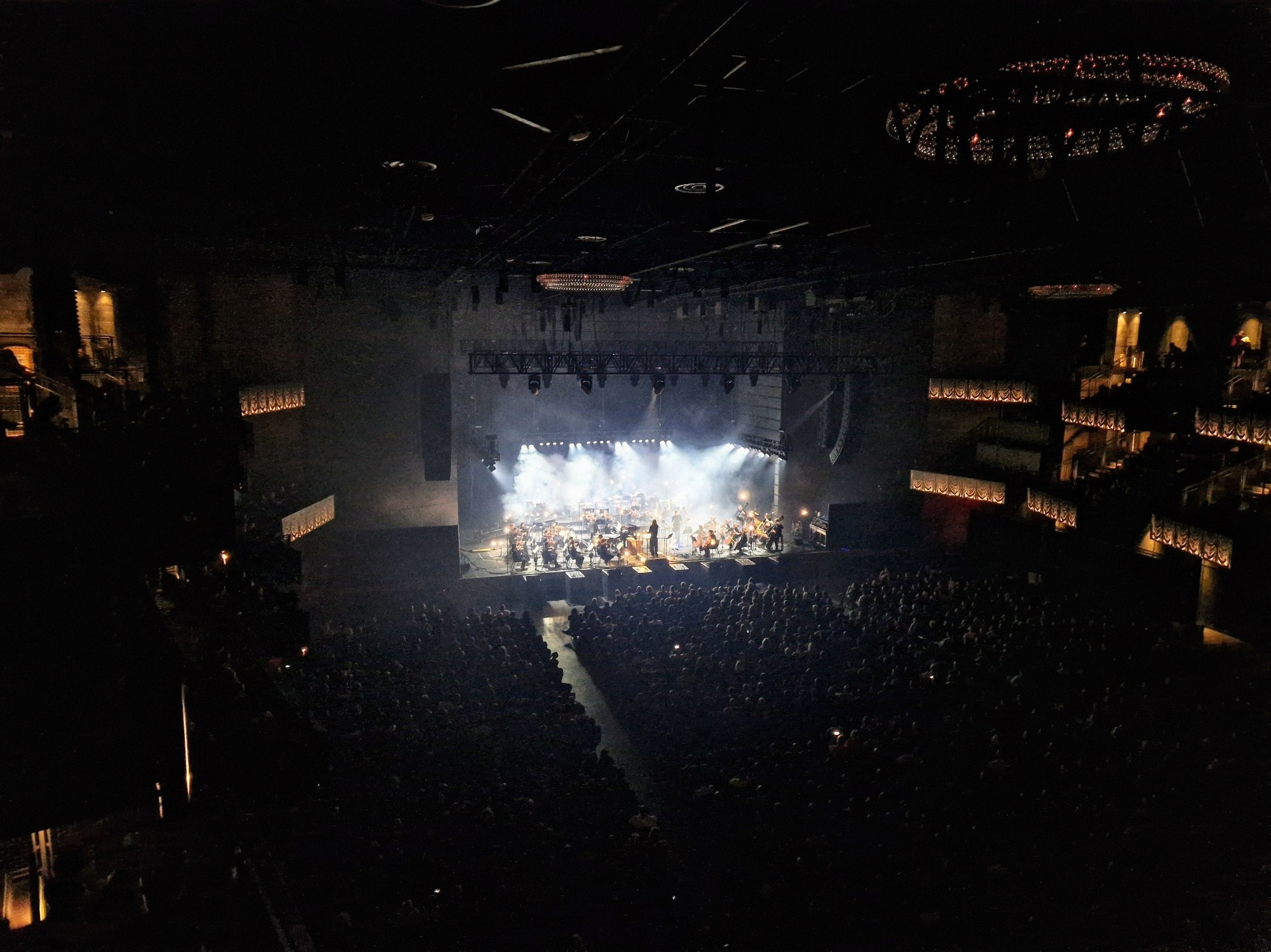 Inside venue. Sigur Rós on stage with an orchestra. Light from stage illuminating venue and audience