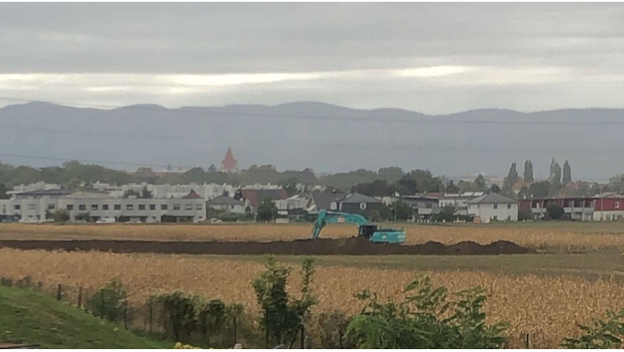 Getreidefeld, in der Mitte ein Bagger, der die Trasse für eine neue Schnellstraße aushebt, im Hintergrund Wiener Neustadt und Gebirge