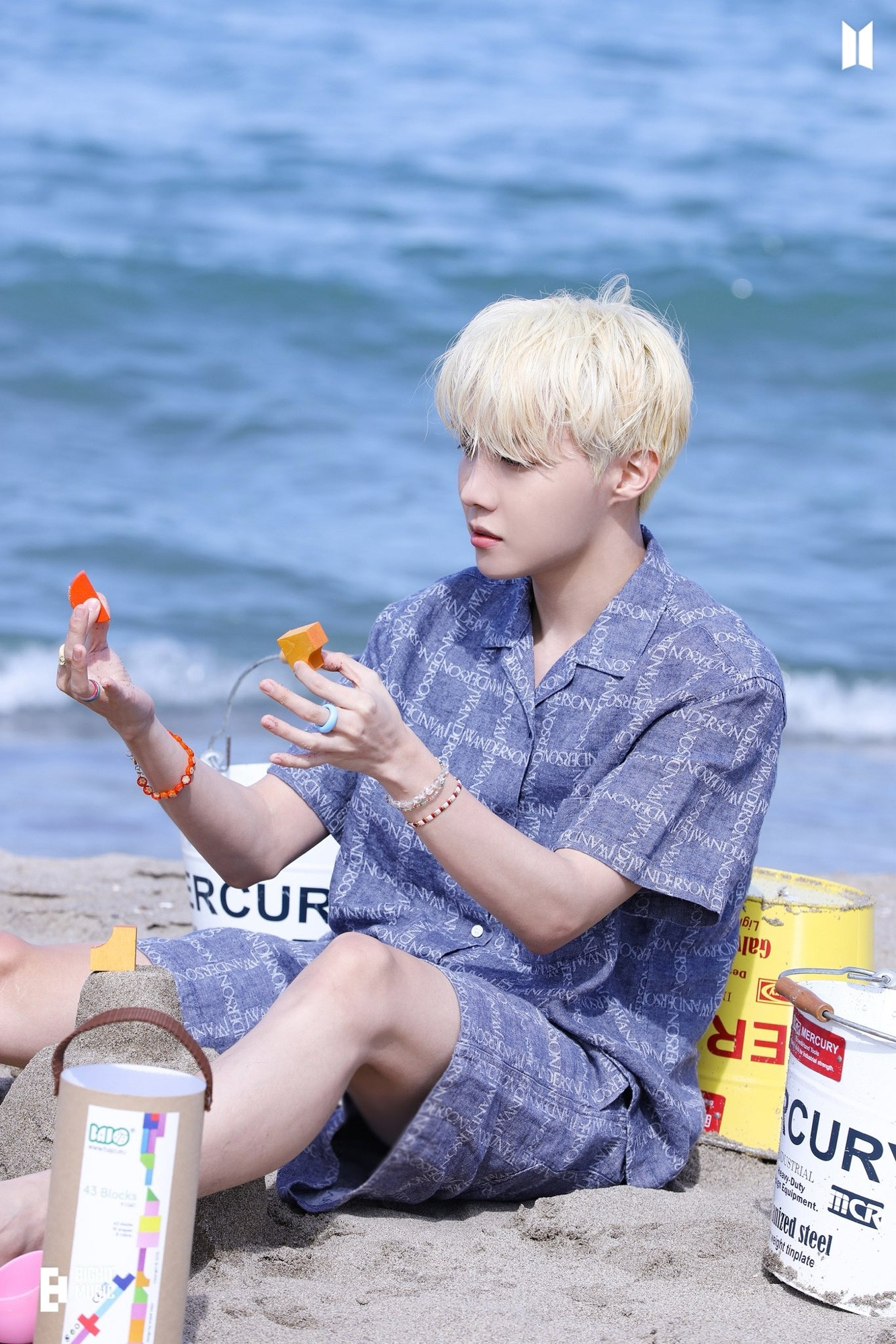 Hobi of BTS sitting on the beach with a blue outfit holding blocks