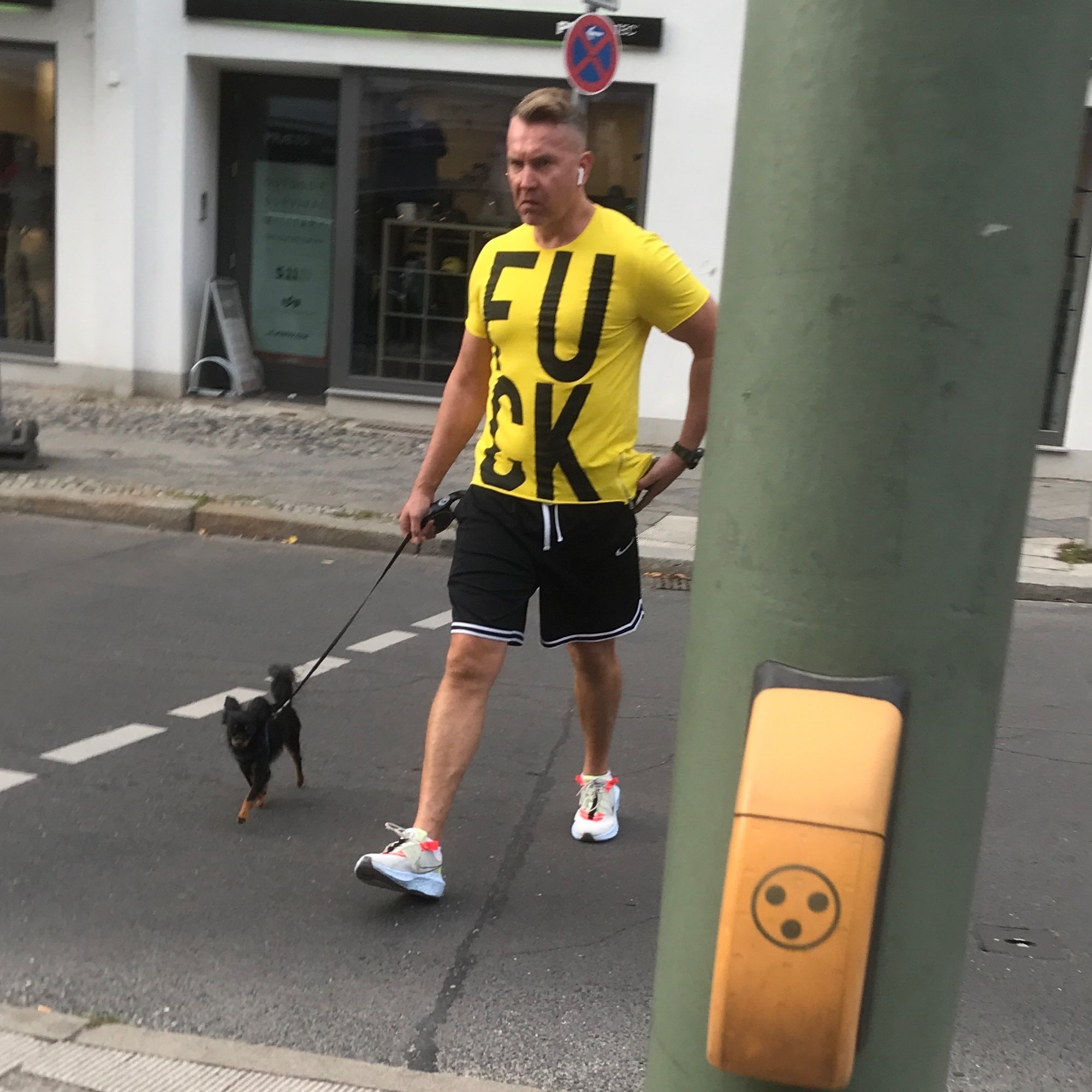 Man wearing earbuds, sneakers, running shorts, and a bright yellow t-shirt with FUCK in large black letters is crossing the street while holding a leash that is attached to a very small dog. In the foreground is a pole with a crossing button.