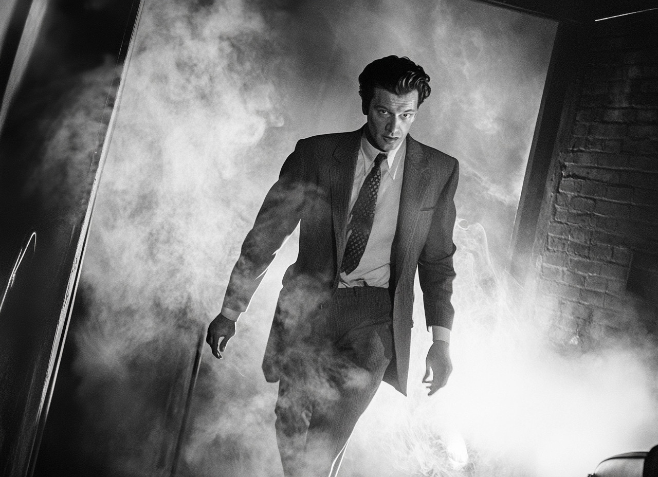 A man in a suit walks through a smoky environment in a black and white photo.