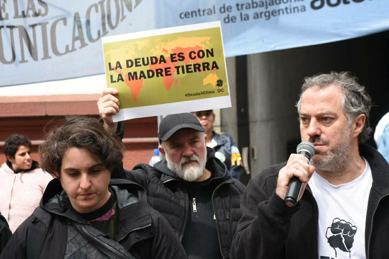 Juan Pablo Olson giving a speech. He wears a debt for climate shirt. Behind him a man holds a placards reading "the debt is with mother earth" in Spanish.