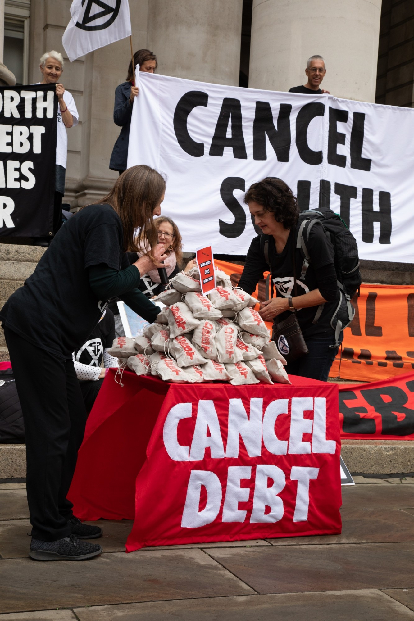 2 people piling up sacks with the debt, that the global north has with the global south. Cancel debt is written on the table cloth. Other debt for climate banners in the background.