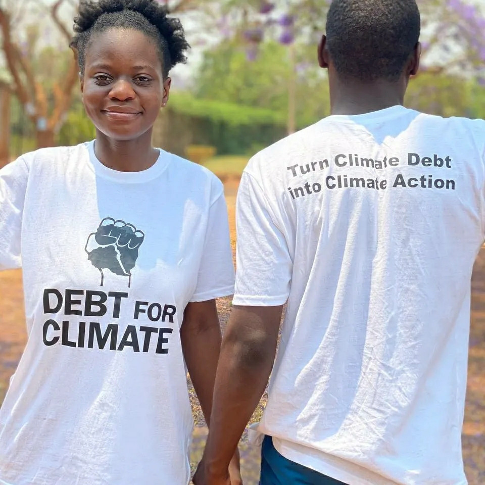 A woman and a man wearing debt for climate shirts. They hold their hands. On his back are the words "Turn Climate debt int climate action"