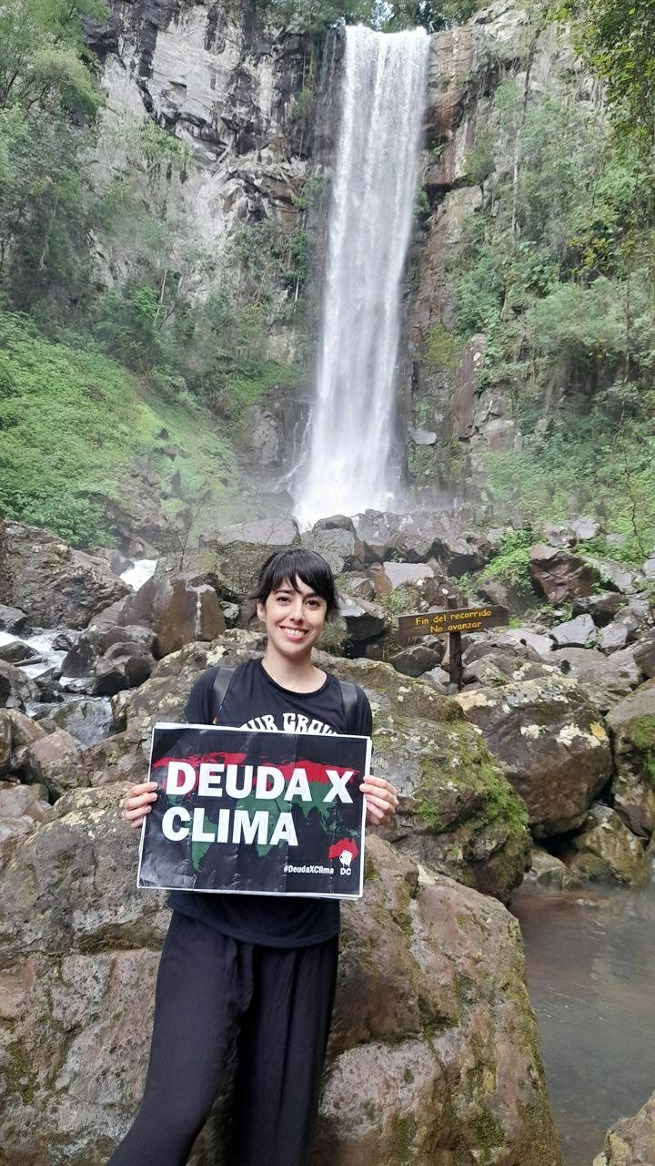 A young woman standing in front of a beautiful waterfall. She holds a Debt For Climate Placard. "Use the Debt for Climate"