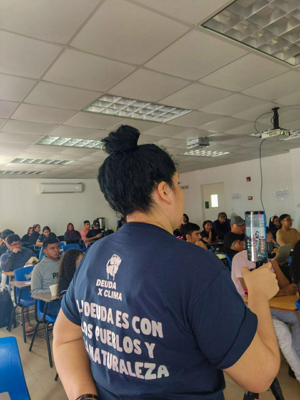 A woman with a debt for climate shirt stands in front of an audience, taking photos.