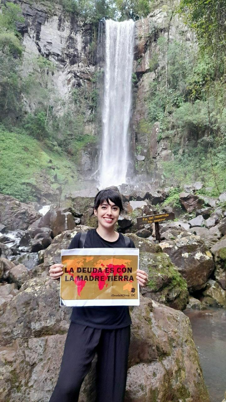 A young woman stands in front of a waterfall. She presents a protest placard smilingly:
The debt is with mother earth.