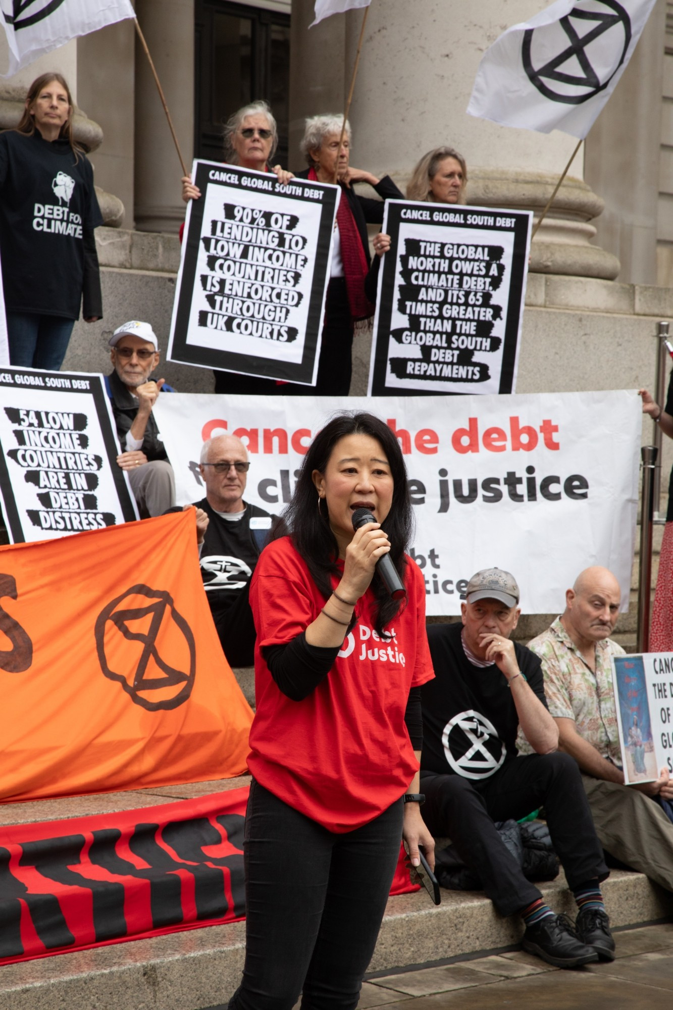 A woman is giving a speech. Debt for climate banners in the background.
Cancel the debt for climate justice. The global north owes a climate debt and it's 65 times greater than the globals debt repayments. A few extinction symbols around.