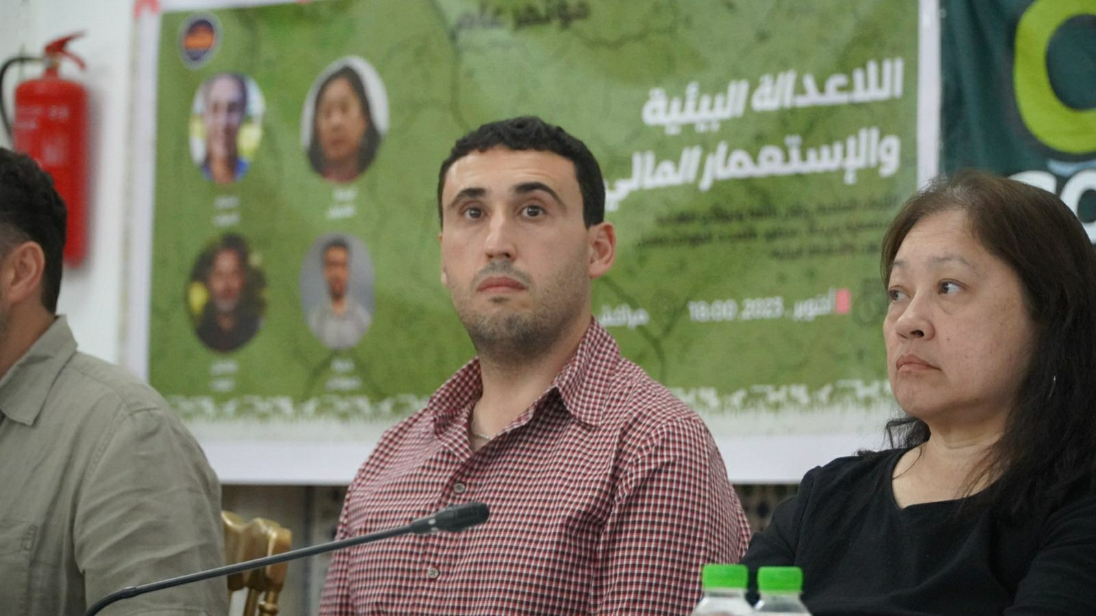 Hamza Hamouchene sitting on the Table next to the other speakers. In the background is a placards presenting the speakers.