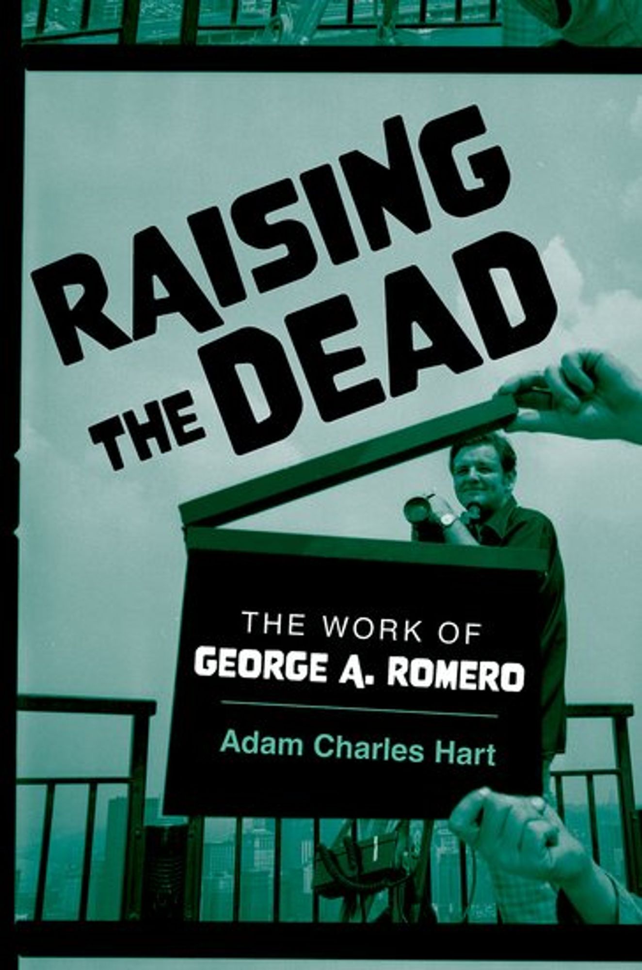 The cover of the book "Raising the Dead: The Work of George A. Romero," in green and black tones, featuring a photo of a young George Romero while sitting on the edge of a tall building, with a film clapboard in front of him bearing the book's title.