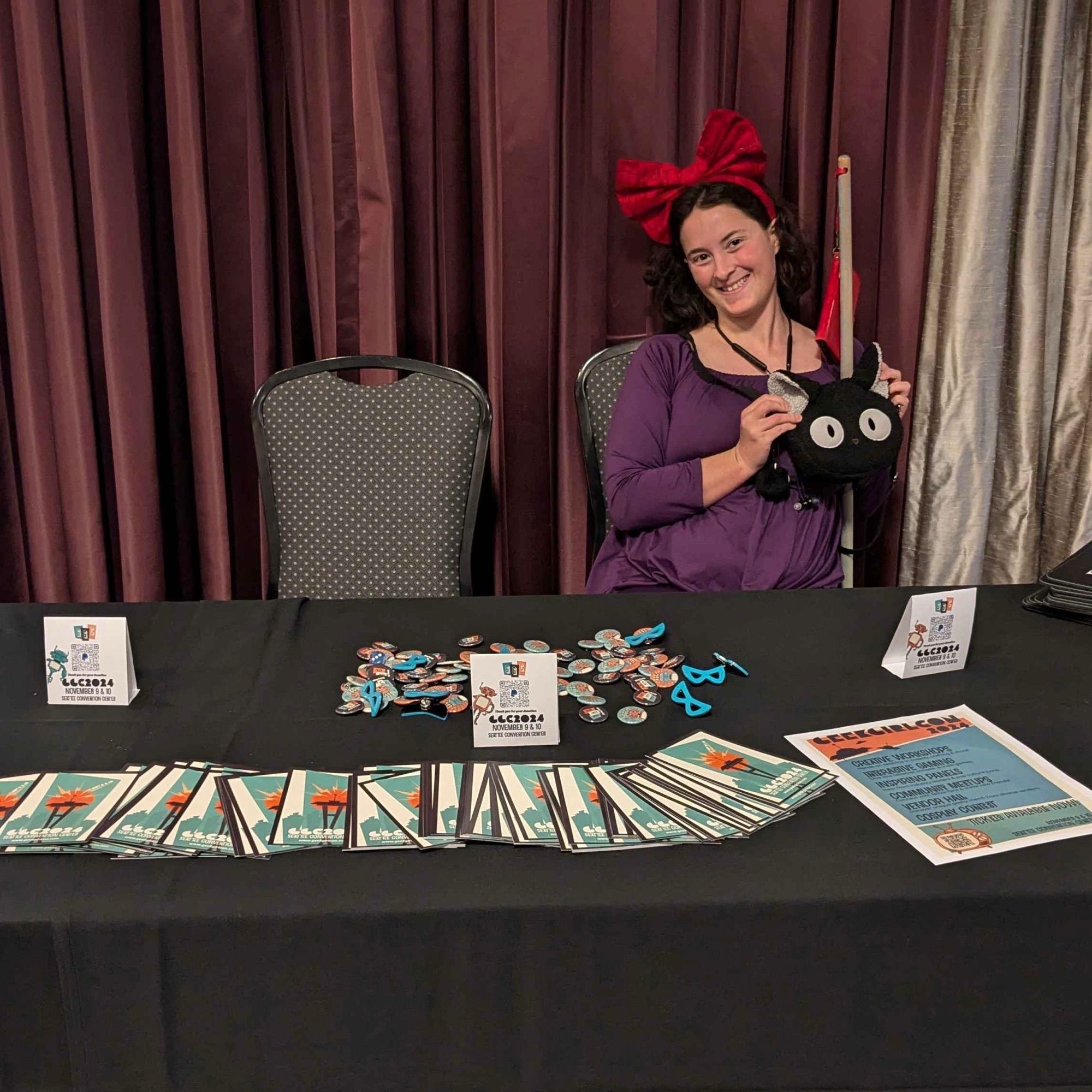 Eli (as Kiki from Kiki’s Delivery Service) sits behind table covered in GeekGirlCon flyers and buttons.